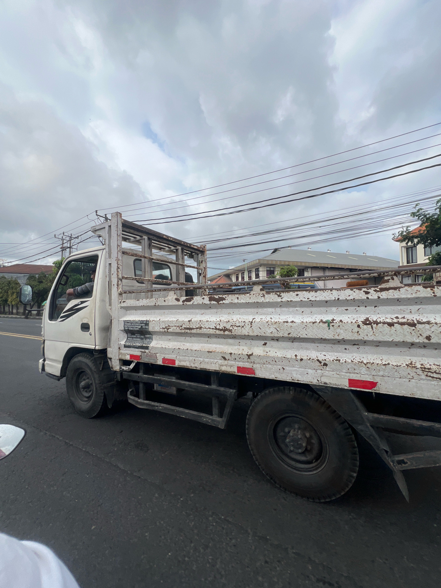 A white truck is driving on a street in Denpasar, Indonesia. The truck is old and rusty, with a metal cage on the back. The driver, a man wearing a striped shirt, is visible through the driver's side window. There are power lines and buildings in the background. The sky is overcast with clouds.