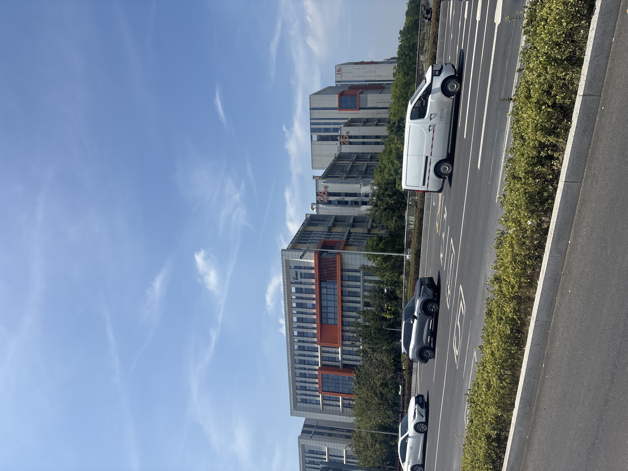 The image depicts a modern street scene in the Suzhou Industrial Park, China, under a clear blue sky with scattered white clouds. In the foreground, a grey SUV and a white sedan are driving on a multi-lane road. Adjacent to the road is a manicured green hedge with a concrete border. Behind the hedge, a line of trees separates the road from a row of contemporary office buildings. The buildings are multi-story structures with a combination of grey, white, and orange facades. Several of the buildings have large orange numerals and Chinese characters visible on their exteriors, indicating numbering, such as "26," "25," and "18." A white panel van is also seen driving on the road, positioned in front of the office buildings. The architecture suggests a commercial or technological district within the industrial park.