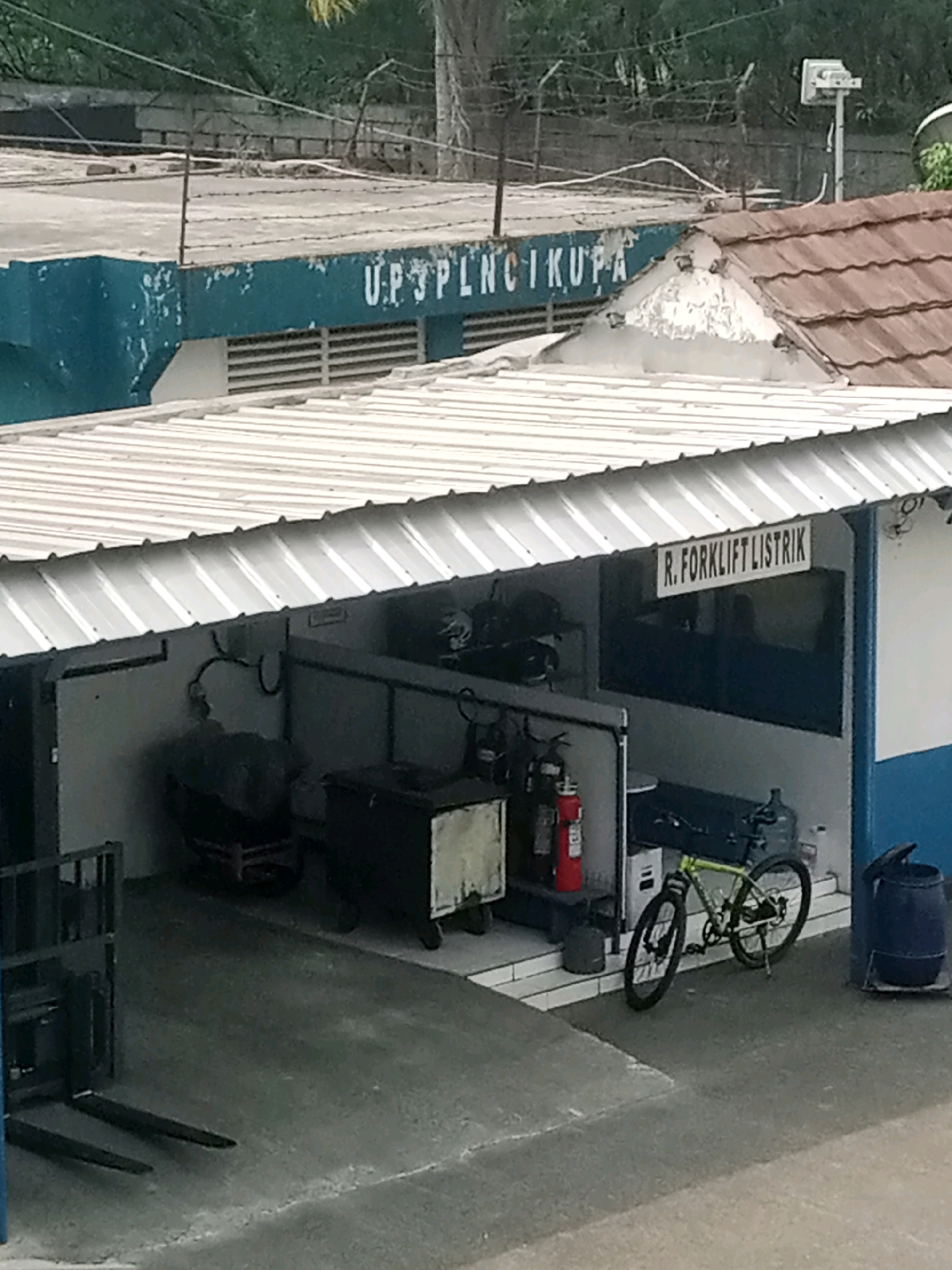 The image shows an exterior view of a building, possibly a depot or workshop, in Balaraja, Indonesia. The building has a blue facade with white lettering that reads "U.P.3PLN CIKUPA" on the upper section. A corrugated metal roof extends from the building, covering an open-air area.
In this covered area, several objects are visible. To the left, a black forklift with two prongs is parked. Adjacent to it, there are what appear to be storage containers or equipment, one of which is light-colored and has a rectangular shape. Further to the right, there is a red fire extinguisher mounted on a stand. A bicycle with a green frame is parked near the center of the scene, leaning against a dark object. To the right of the bicycle, a dark blue barrel with a lid is situated.
The background reveals a fence with barbed wire, suggesting a secure perimeter. Trees and vegetation are visible behind the fence. The ground surface in the foreground is paved, likely concrete. The overall scene depicts a utilitarian environment, possibly related to utility services, given the lettering on the building. No people are clearly visible in the image.