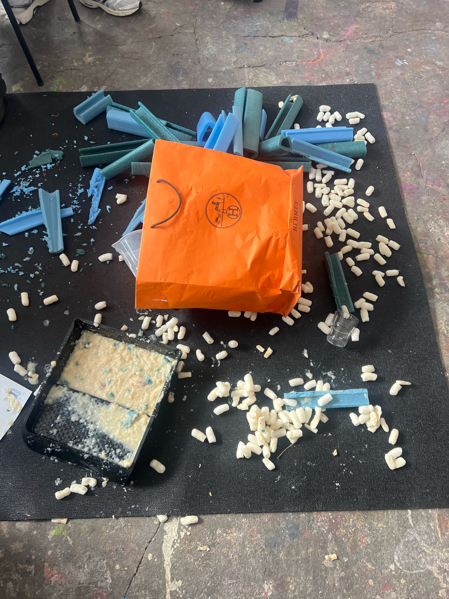 An overhead, medium shot captures a chaotic, art-studio floor in Berlin, Germany, littered with the remnants of an unboxing. The scene is dominated by a large, black mat, likely a protective surface, spread across a concrete floor splattered with colorful paint.
In the center of the mat, an orange Hermès shopping bag lies open, its contents spilled out. Blue and green foam packaging pieces, some curved and cylindrical, others angular and broken, are scattered around the bag. A small, clear glass with a green foam wrapper rests near the bag, and a black plastic tray filled with a creamy, off-white substance with blue flecks is positioned to the left. Numerous small, white, peanut-shaped packing peanuts are strewn across the mat, suggesting a hasty unboxing.
The background shows a portion of a grey concrete floor with colorful paint splatters. The general impression is one of a creative space disrupted, possibly during or after a delivery.
