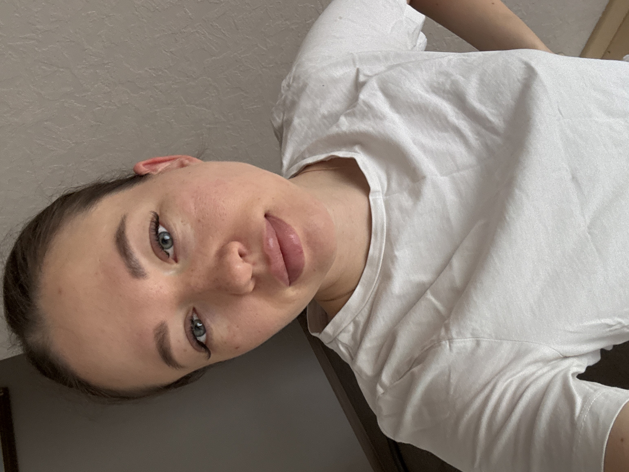 A woman is posing indoors for a selfie, with her face slightly turned to the side. She has light skin, blue eyes, and dark hair pulled back. She is wearing a white T-shirt. The background is a neutral wall, with a door frame visible in the lower left corner. The image provides no information about the location in the city of Ферма, Russia.