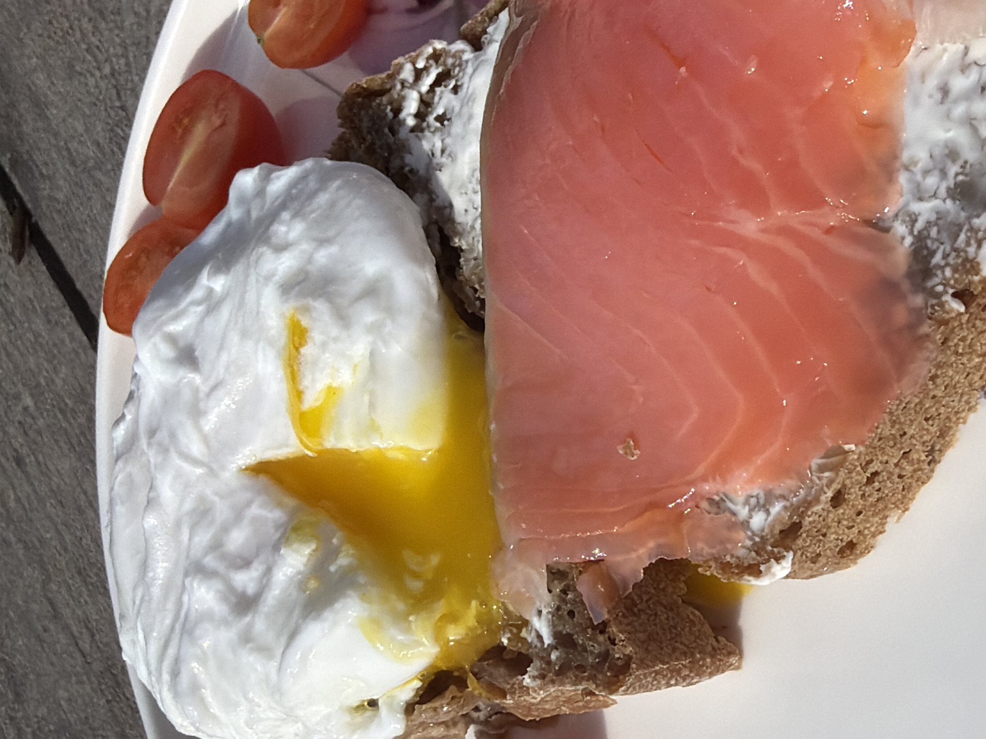 A close-up shot features a plate containing food. Visible are slices of tomato, a poached egg with a runny yolk, and a piece of bread topped with cream cheese and a slice of salmon. The plate appears to be on a wooden surface. The image does not contain information that would indicate the location as Chaikovsky, Russia.