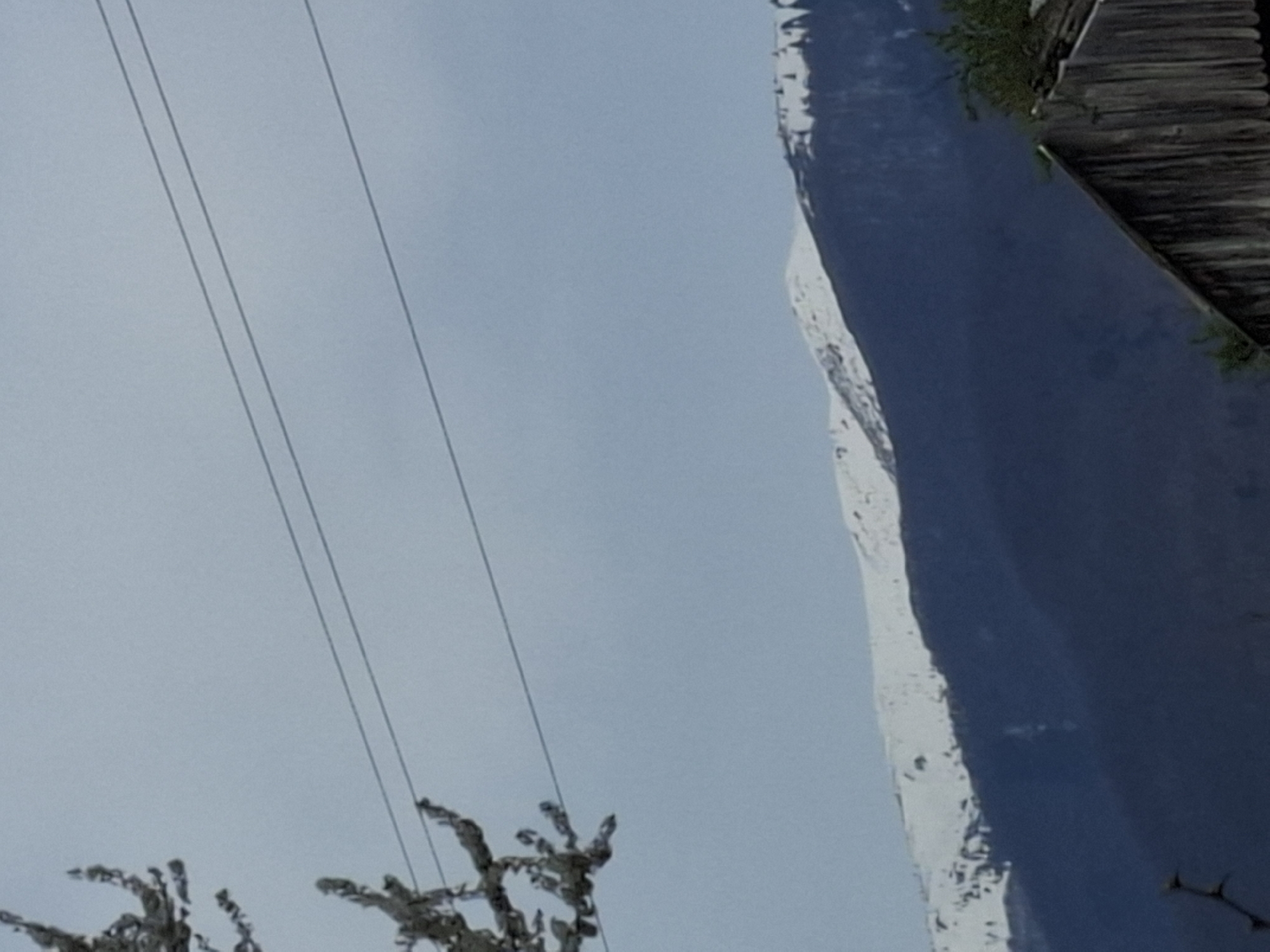 The image, possibly taken in Barziya, Bulgaria, shows a wintry scene. Power lines cut across a pale, overcast sky. Silhouetted, possibly snow-covered trees are visible in the lower left. The right portion of the frame is dominated by what appears to be a snow-covered mountain slope with a dark blue lake or river at its base. A portion of a wooden structure, possibly a building, is situated above the lake.