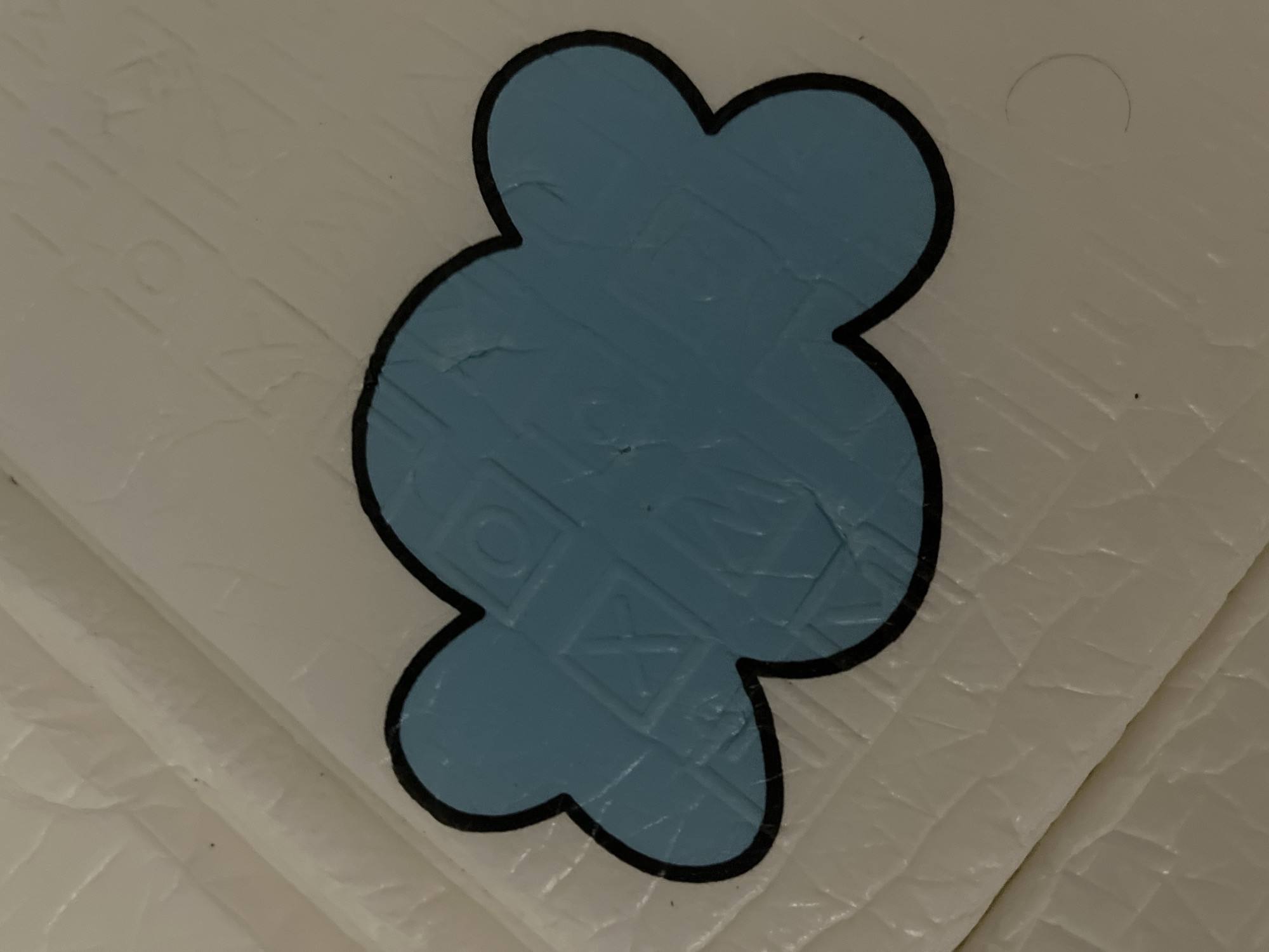 A blue cloud-shaped sticker with a black outline is placed on a textured white surface. The white surface has a diamond-shaped pattern embossed on it. The sticker appears to be slightly creased. No people or other objects are visible. The location is not identifiable from the image.