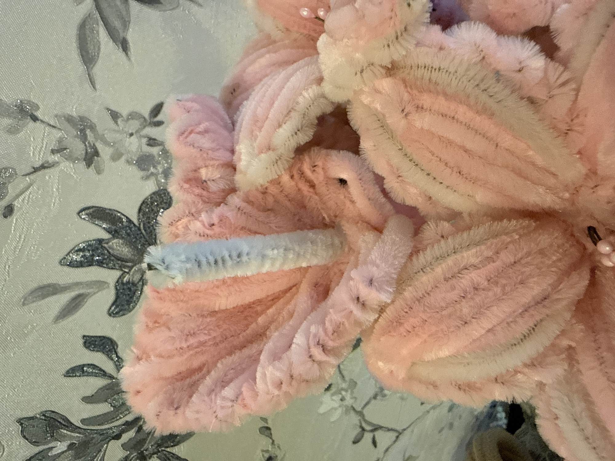A close-up shot features a fluffy pink floral decoration against a backdrop of patterned wallpaper with silver and gray floral designs. A light blue and white pipe cleaner-like piece is incorporated into one of the petals of the pink floral object. The photograph appears to be taken indoors, with an emphasis on texture and detail.