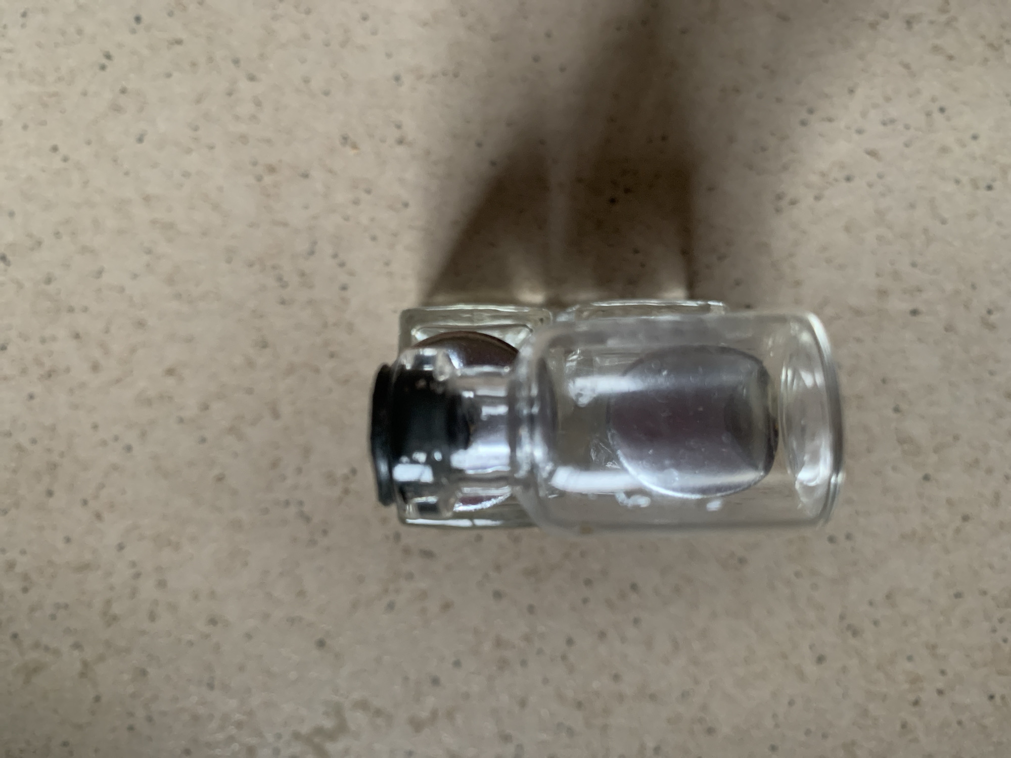 A small, clear glass vial with a black stopper sits on a speckled, beige surface. The vial has a rectangular base and a round opening at the top. Inside the vial, a translucent, purplish object is visible. A shadow cast by the vial and stopper is present on the surface to the right.
