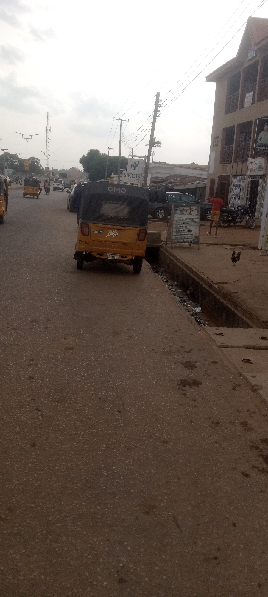 The image shows a street scene in Sabon Tasha, Nigeria. A yellow tricycle with black covering is positioned in the middle of the street, facing away from the viewer. A sign with "OMO" is visible on the covering. Other vehicles and people are on the street, including other tricycles and a person standing near a building on the right side of the image. A chicken stands on the sidewalk to the right of the tricycle. Buildings are visible in the background. Power lines stretch across the sky. A sign is displayed by the side of the road with the words "SIKOJS PHARMACY." A storm drain runs alongside the road.