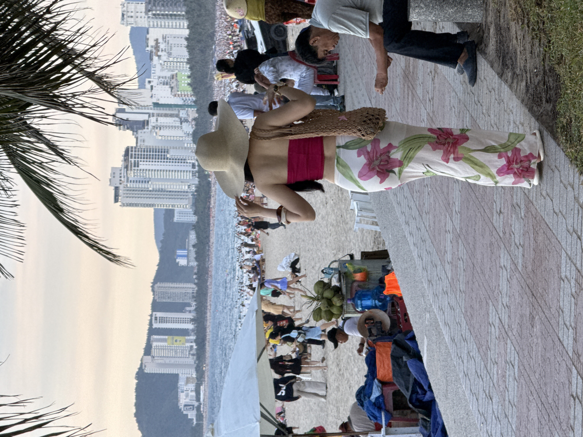 In Nha Trang Ward, Vietnam, a busy beach scene unfolds with a backdrop of high-rise buildings. In the foreground, a woman in a wide-brimmed straw hat and a floral-patterned skirt walks away from the camera, carrying a woven bag. To her right, a man in a light gray shirt and dark pants sits on a stone bench, looking down. Behind them, a crowded beach stretches towards the ocean, dotted with numerous people. Various beach umbrellas and stalls are visible on the sand, along with a collection of coconuts and bottles on a cart. The sky is overcast, suggesting either early morning or late afternoon.