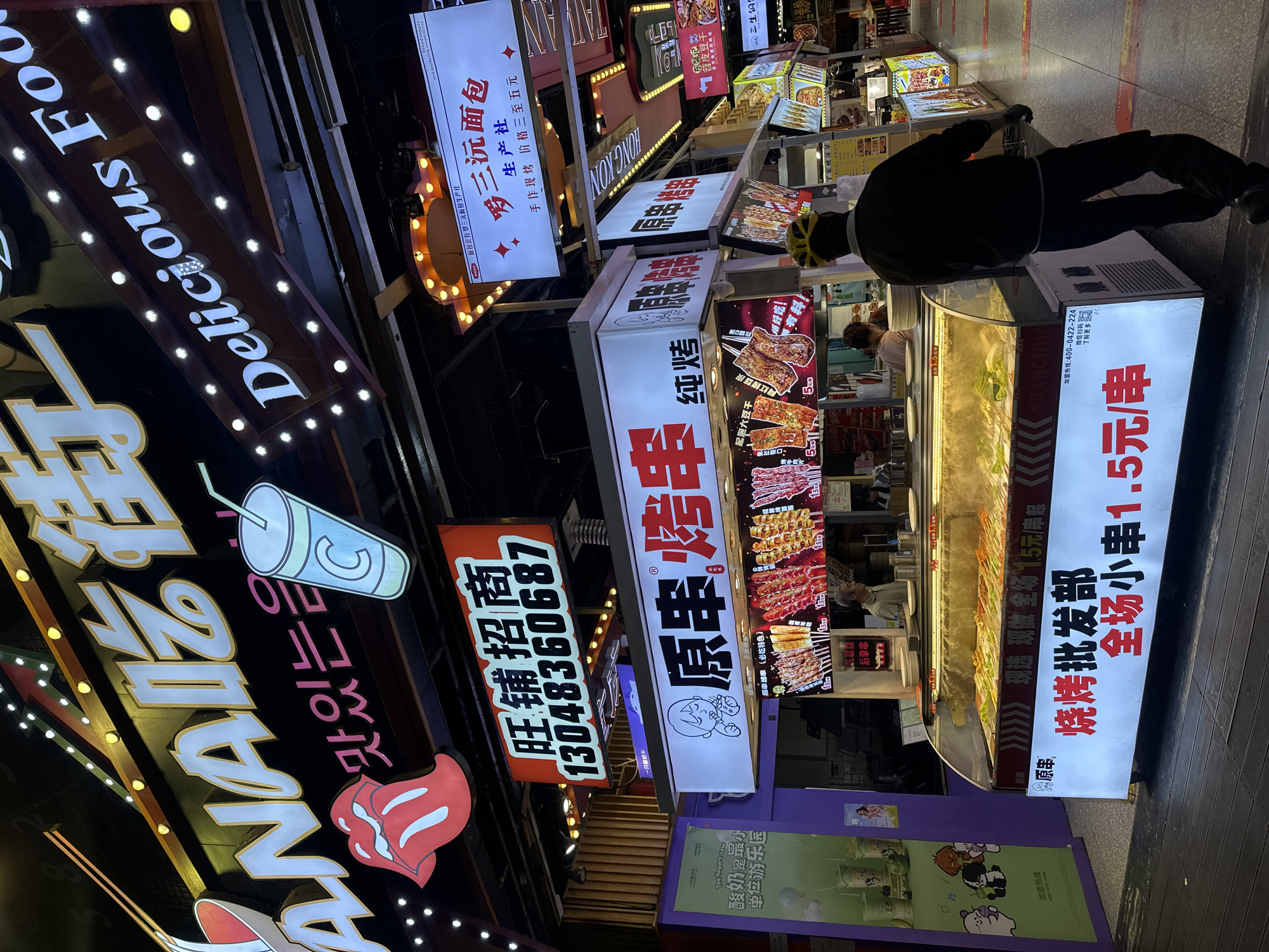The image shows a vibrant night scene at a street food market in Chongqing High-tech Zone, China. The foreground is dominated by a food stall displaying skewers of various meats and vegetables under bright lights. A person wearing a yellow helmet and dark clothing is walking away from the stall towards the right. Above the stall, prominent signage in Chinese characters advertises "原串" (Original Skewers) and "烤串" (Grilled Skewers), with prices displayed for "全场小串" (all small skewers) at 1.5 Yuan per skewer.
To the left of the food stall, a purple pillar is visible with signage for "The Yogurt-cow" and cartoon characters. Further up, a large illuminated sign reads "ANANZ" in white letters with a red lips graphic, and below it, Korean text that translates to "delicious food". To the right, a sign with white text on a brown background says "Delicious Food" with illuminated bulbs. Another sign above this reads "三三沉面包" (Sansan Sinking Bread).
The environment is characteristic of a busy night market with multiple food vendors, neon lights, and signage creating a lively atmosphere. The overall impression is one of a bustling commercial area focused on street food.