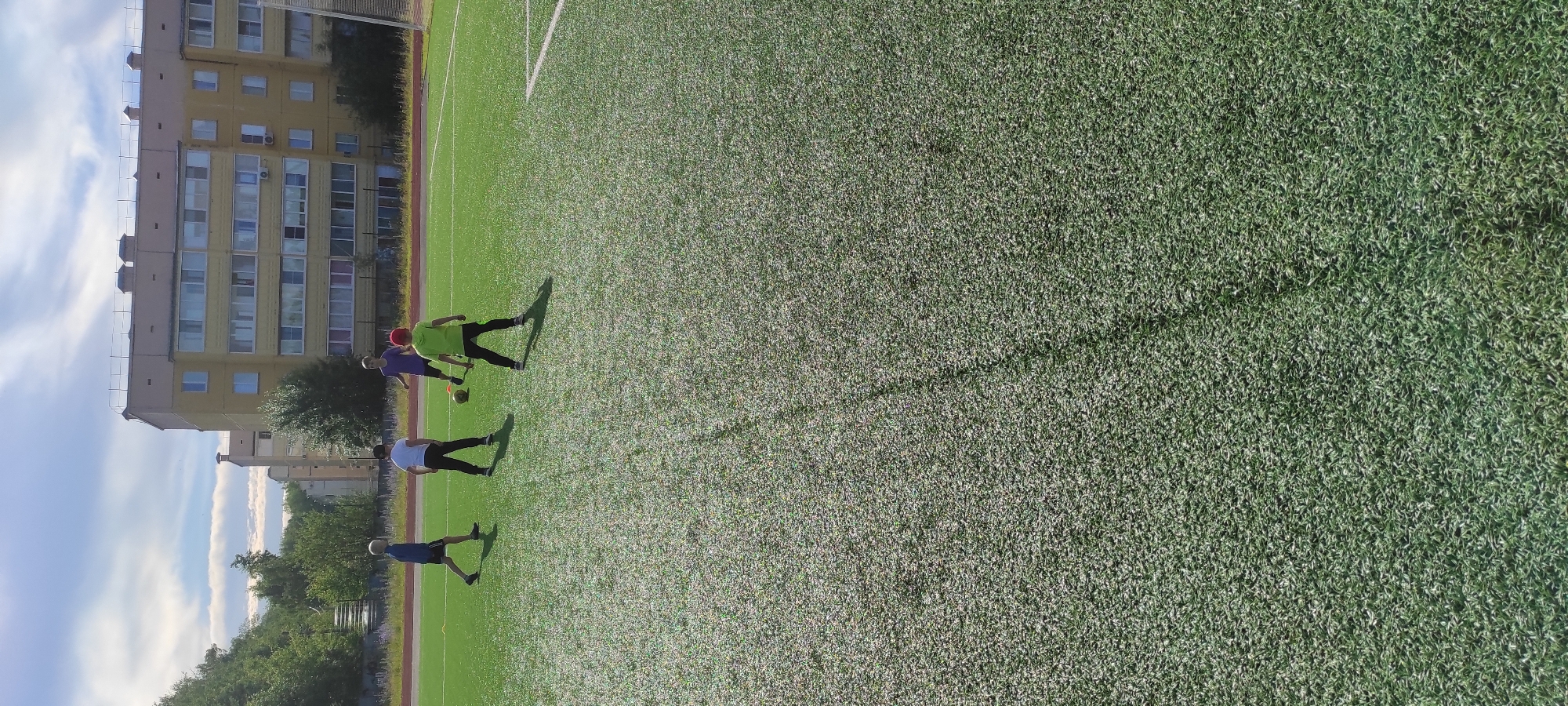 Four children are playing on a green artificial turf soccer field. The field is marked with white lines. In the background, a multi-story yellow apartment building with many windows is visible, along with trees and a cloudy blue sky. The children are dressed in athletic clothing. One child on the left wears a blue shirt and dark shorts. Another child in the center left wears a white t-shirt and dark pants. A child in the center right wears a purple shirt and dark pants, and is near a soccer ball. The fourth child on the right wears a green shirt and dark pants. The scene appears to be a sunny day.