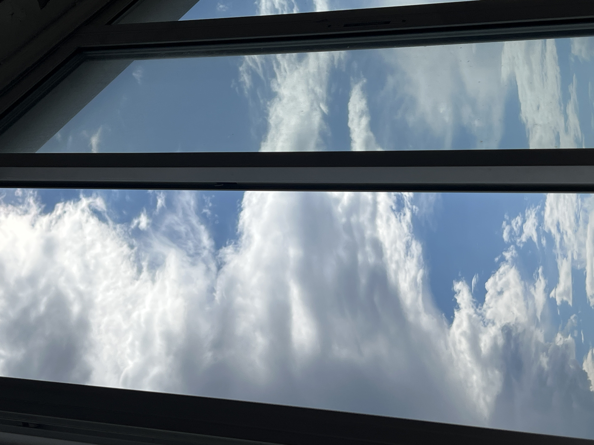 The image is a view of a cloudy sky through a window frame. The window appears to be part of a modern building, with dark gray metal framing. The sky is a mix of blue sky and white and gray clouds, with some areas of bright sunlight peeking through. There are no people or any discernable objects in the foreground. The scene depicts a natural, outdoor environment viewed from an indoor perspective, and does not contain any information that would suggest its location within the city of Tolyatti, Russia.