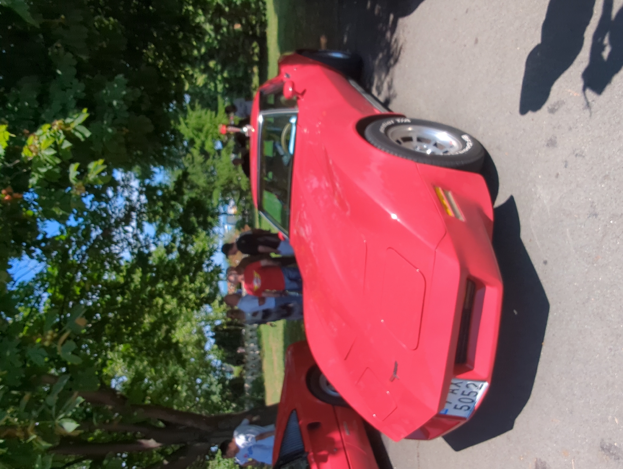 An outdoor scene shows a red Chevrolet Corvette C3 parked on asphalt. In the background, several people are gathered under the shade of leafy trees. A license plate on the front of the Corvette reads "AX 5052". The setting appears to be a car show or gathering, possibly in the city of Burgas, Bulgaria, given the license plate format. The sky is visible through the trees, suggesting a sunny day.