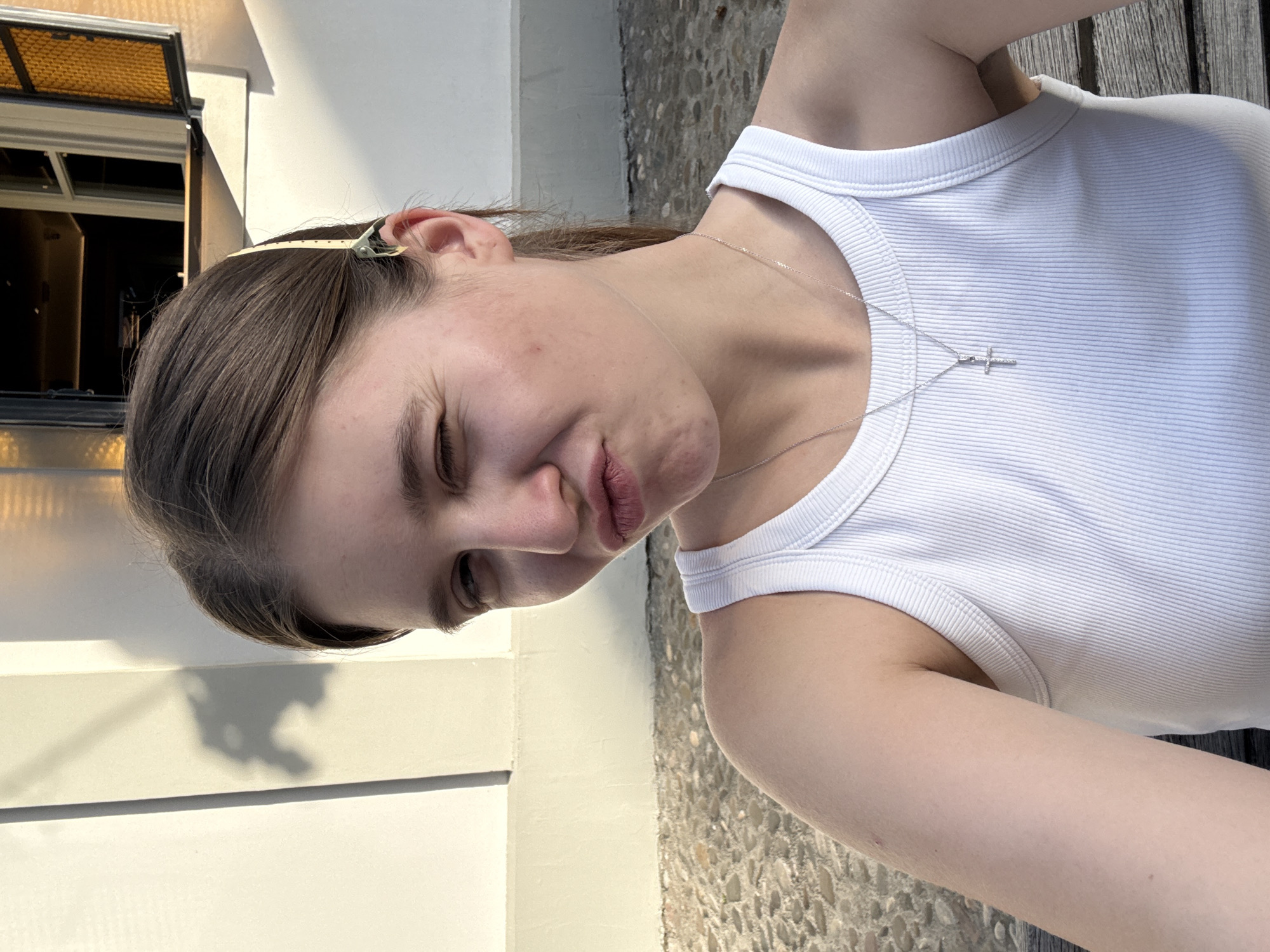 A young woman is depicted in a medium shot, partially angled. She is wearing a white ribbed tank top and a delicate necklace with a cross pendant. Her hair is brown and partly held back with a clip. A window is slightly visible in the background, reflecting sunlight. The background includes a white wall and stone or concrete flooring. The location is indicated as Belgrade, Serbia, inferred from visual features in the background potentially common to the city.