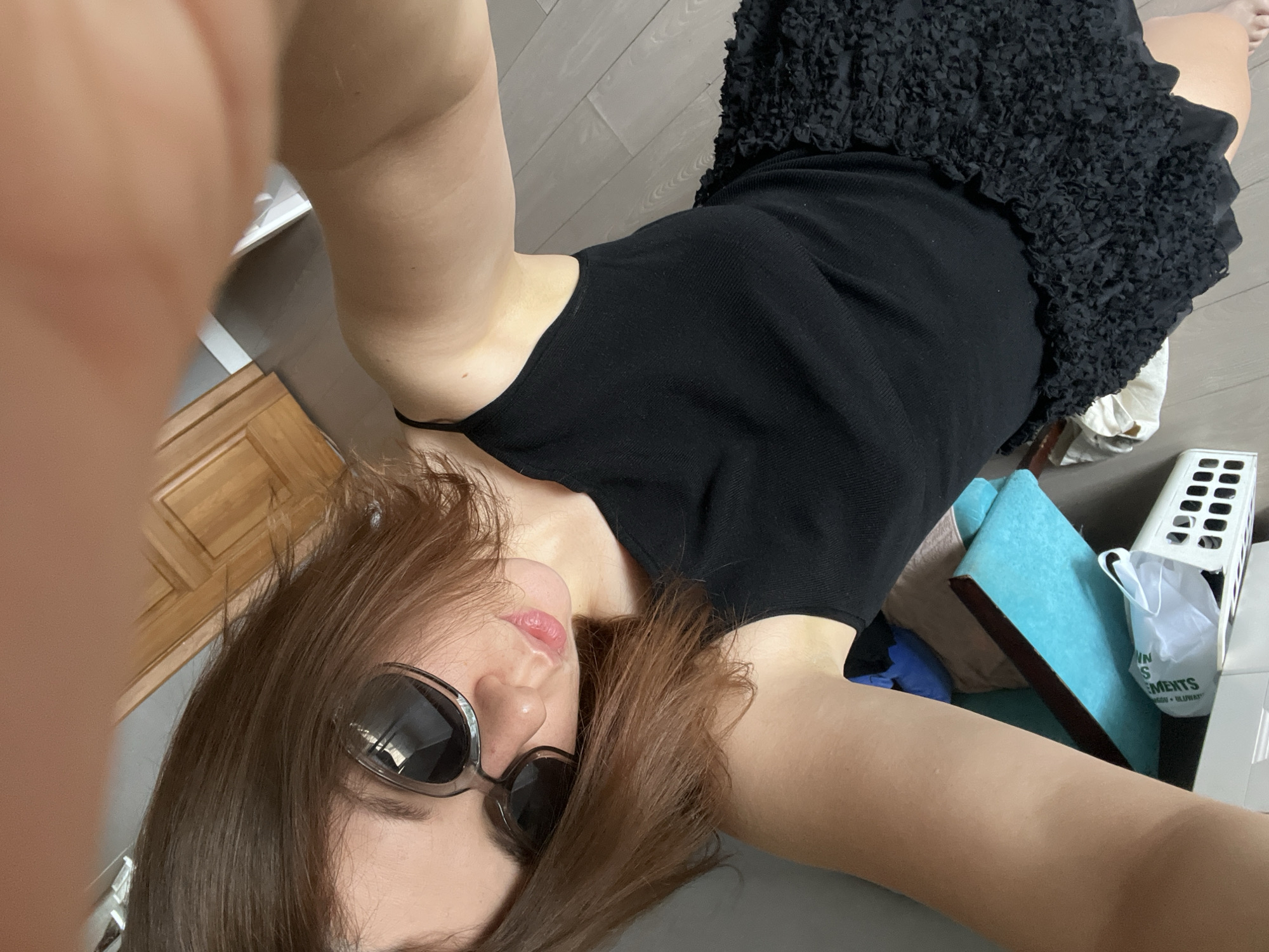 A young woman with brown hair and light skin, wearing sunglasses, a black tank top, and a black ruffled skirt, is lying on the floor and taking a selfie. The background shows a wooden floor, a wooden door, and some objects on the floor, including a blue book and a white plastic basket. The scene appears to be indoors, likely in a residential setting. The image does not provide any information about the location in Krasnoyarsk, Russia.