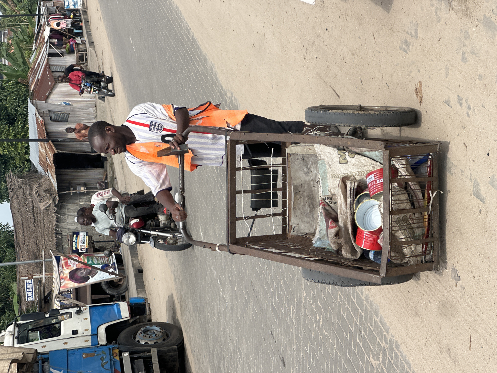 An outdoor, eye-level, medium shot captures a man pushing a rusty, two-wheeled cart filled with various items on a paved street in Badagry, Nigeria. The man, wearing a white and red striped shirt with an orange vest, black pants, and a ring on his left ring finger, looks to his left. The cart contains a bag, several red cans, and other unidentifiable objects. In the background, a large white and blue truck is parked on the left. To the right of the truck, two men are riding a motorcycle. Further in the background, a row of buildings with corrugated metal roofs and wooden structures lines the street. Trees and foliage are visible behind the buildings. The ground is a mix of pavement and dirt. A sign with "REV. OKE TOJINU STR." is visible on a pole in the background.