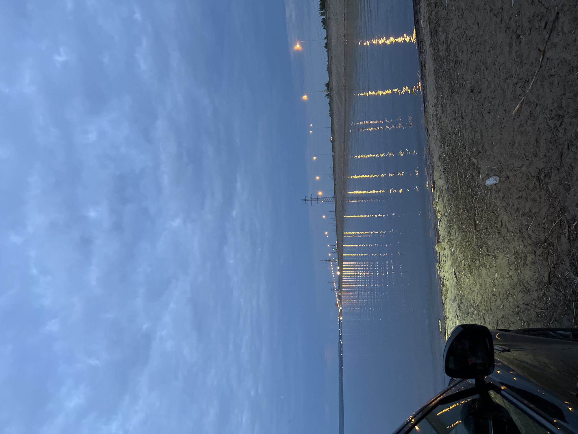 The image depicts a twilight scene along the coast of Noviy, Russia. A black car is parked on a sandy embankment, with its headlights illuminating the immediate foreground and the water's edge. The body of the car and its side mirror are visible on the left side of the frame. The water, likely a bay or large river, reflects the lights of streetlamps and utility poles that line a distant causeway or road. These lights create a shimmering, elongated trail on the water's surface. The sky is overcast with grey clouds, typical of evening or a partly cloudy day. The overall impression is one of a quiet, natural setting enhanced by man-made illumination.