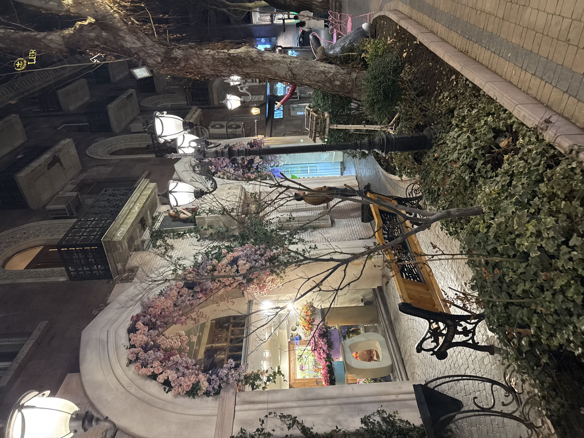 The scene depicts a well-lit urban street at night in Yerevan, Armenia. The dominant structure on the left is a multi-story building featuring a prominent ground-floor shop entrance styled as a large archway, heavily adorned with artificial pink and purple floral garlands. Inside the brightly illuminated shop window, various potted flowers in vibrant pink, purple, orange, and yellow are displayed on pedestals and in baskets, alongside a white, sculptural object. Above the shop, the building exhibits multiple dark balconies with black metal railings and arched windows.
A classical white street lamp is mounted on the building's upper left. A tall, ornate black lamppost with three frosted glass lanterns stands in the mid-ground. To its right, a mature tree with rough, light-brown bark and mostly bare branches occupies a significant portion of the right foreground. A smaller, bare sapling is positioned closer to the lamppost. The pavement consists of two-toned interlocking bricks, forming a pedestrian path.
A raised planter bed separates the building from the path, filled with low-lying green ivy and sparse shrubs. A wooden park bench with black ornate metal legs, bearing the inscription "www.yerevan.am", is positioned in front of the planter. Near the large tree, a small wooden bird feeder on a stand is visible, along with a dark statue of a rabbit or hare accompanied by two pink wire-frame chairs. Higher on the building's facade, a small figurine of a woman in a red garment leans from a decorative alcove.
One male individual in a brown jacket stands near the shop entrance, facing inwards. Further down the street on the right, two additional individuals are visible walking away from the camera, one wearing a white coat. Overhead, illuminated signs, including a golden "LER" with a fleur-de-lis, are visible against the dark facades of distant buildings. Air conditioning units are present on some balconies of the background buildings.