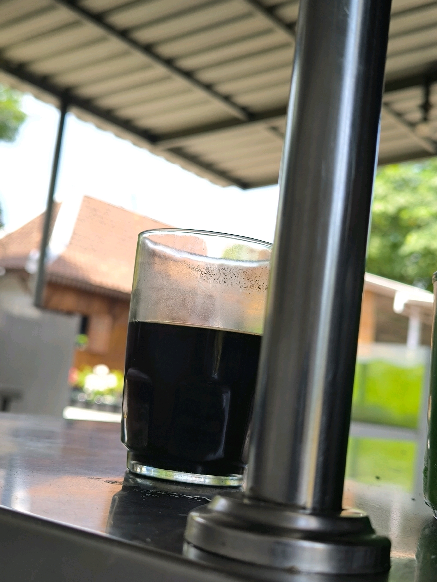 A glass of dark liquid, likely coffee, sits on a metallic surface in the foreground. A thick, metallic pole obstructs the view to the right. In the blurred background, a building with a terracotta roof is visible, along with some greenery and sunlight filtering through an overhead structure. The scene appears to be outdoors, possibly on a patio or balcony, with a slightly overcast sky.