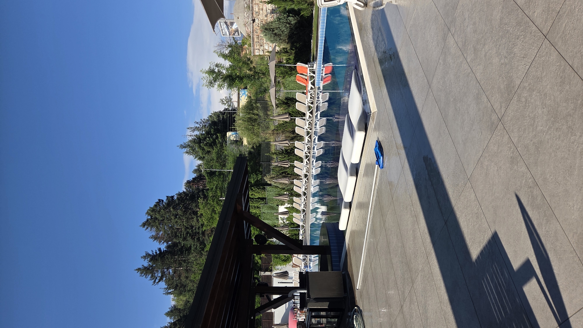 The image depicts a poolside scene at a resort in Bania, Bulgaria. The foreground is dominated by a tiled patio with a long shadow cast by a structure, possibly a pergola or canopy. A blue cleaning tool is visible on the patio. Beyond the patio, a large swimming pool is the central focus. Several submerged lounge chairs are positioned within the pool, offering a unique seating experience. The pool is surrounded by a row of white lounge chairs and umbrellas, suggesting a relaxation area for guests.
In the background, a lush green hillside covered with trees rises behind the pool. Some buildings and structures are visible nestled among the trees. The sky is a clear, vibrant blue, indicating good weather. The overall impression is one of a well-maintained outdoor leisure facility. The presence of a bar area with stools and bottles to the left suggests an area for refreshments. The stone wall structure on the right side of the image hints at a traditional architectural element incorporated into the resort's design. The clear blue sky and abundant greenery point to a location that likely experiences warm and pleasant weather.