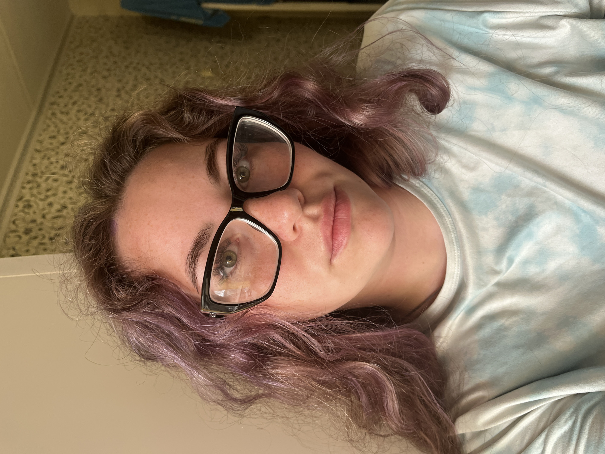 A woman with purple hair wearing black glasses is lying on a surface, possibly a bed or a floor. She is wearing a tie-dye shirt of light blue and white. The background shows a tan, patterned floor and a white wall. A blue object is visible out of focus in the upper center of the frame. The scene is brightly lit. The location is not discernible from the image.