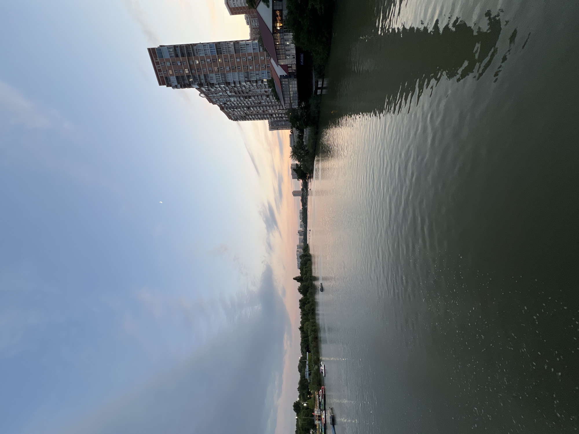 The image captures a scenic view of the Kuban River in Krasnodar, Russia, during a twilight hour, likely just after sunset. The sky displays a gradient of soft blues and pinks, with a crescent moon visible high up. Wispy clouds stretch across the horizon.
On the right side of the frame, a tall, modern apartment building, characteristic of urban development, stands prominently. Its brick facade and numerous windows suggest a residential area. Adjacent to it, a lower building with a red-tiled roof and large glass windows, possibly a cafe or restaurant, is visible. A sign with Cyrillic lettering, "КАФЕ" (KAFE), is discernable on this structure.
The river itself occupies the majority of the foreground, its surface rippling gently, reflecting the colors of the sky and the surrounding architecture. Several small boats are scattered across the water, indicating recreational activity. On the left bank of the river, a stretch of green trees and vegetation lines the shore, interspersed with lights that suggest a populated area or a waterfront park. Further in the distance, the city skyline is visible with more buildings.
The overall atmosphere is tranquil and serene, highlighting the coexistence of nature and urban life.