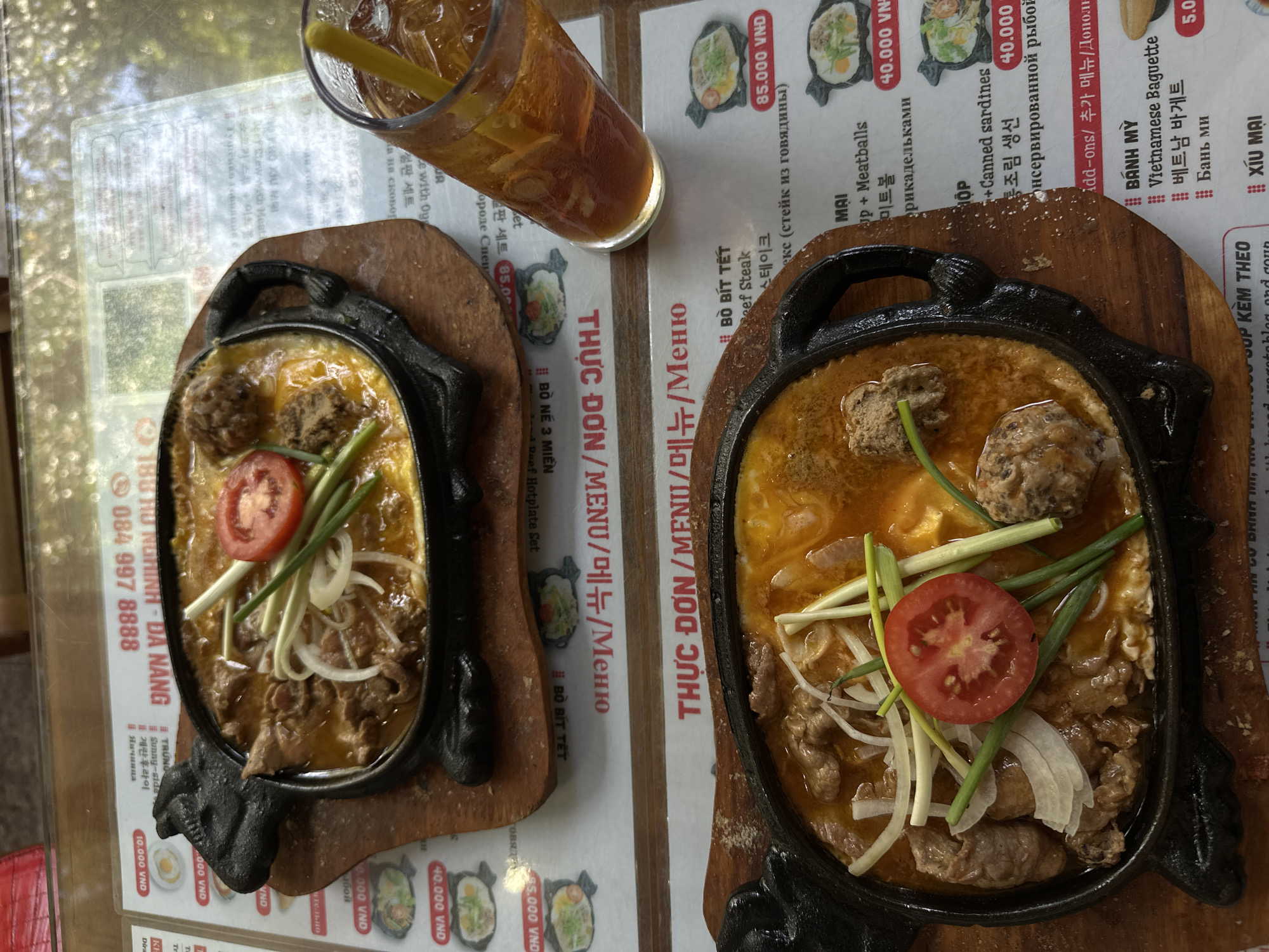 The image shows two sizzling hot plates of food and a glass of iced tea on a table in a restaurant in Đà Nẵng, Vietnam. The hot plates contain meat, meatballs, a sliced tomato, and green onions served with a side of what appears to be scrambled eggs. The dishes are presented on wooden stands. The restaurant's menu is visible beneath the hot plates, featuring text in Vietnamese, English, Korean, and Russian, indicating an international clientele. One menu item visible is "Bò bít tết Beef Steak" for 85,000 VND. The overall environment suggests a casual dining establishment.