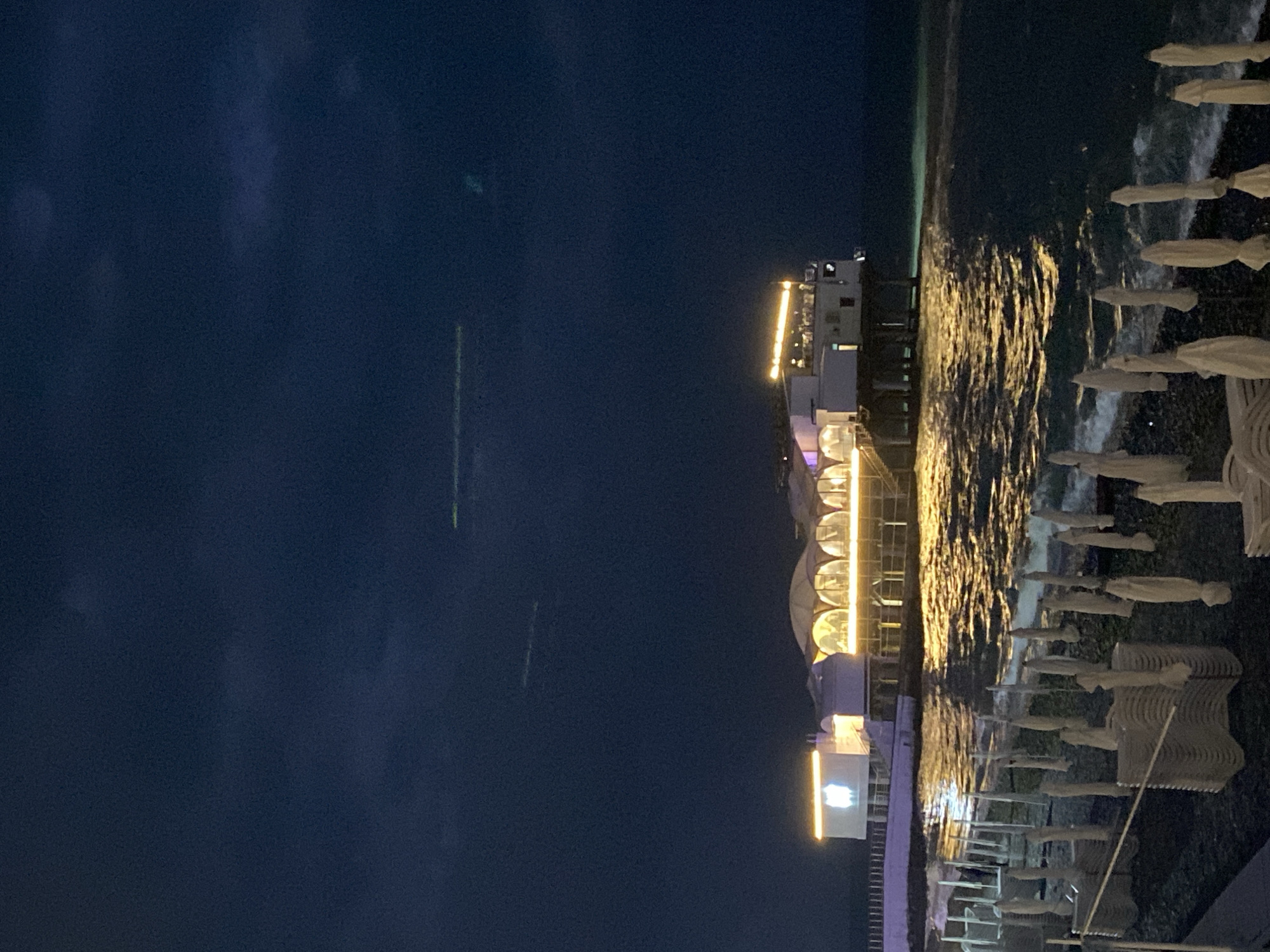The image depicts the beach in Sochi, Russia, at night. A pier illuminated with bright yellow lights extends out into the dark sea. The beach is lined with closed, white sun umbrellas, and there are several white beach chairs stacked in rows. The sky is dark and cloudy. The sea is reflecting the light from the pier and the nearby buildings.