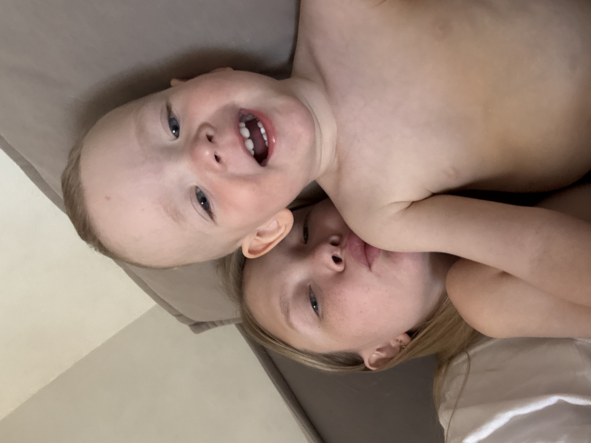 In a close-up, indoor shot, a young Caucasian child with blonde hair and blue eyes is the primary subject, smiling with their mouth open and teeth visible. To the left and slightly behind, a young Caucasian female with blonde hair and fair skin is visible, puckering their lips as if to kiss the child. Both individuals appear to be on a bed or couch with a neutral-colored headboard. The lighting suggests an ambient indoor setting. The specific location in Chaikovsky, Russia, cannot be determined from the visual information provided in the image.