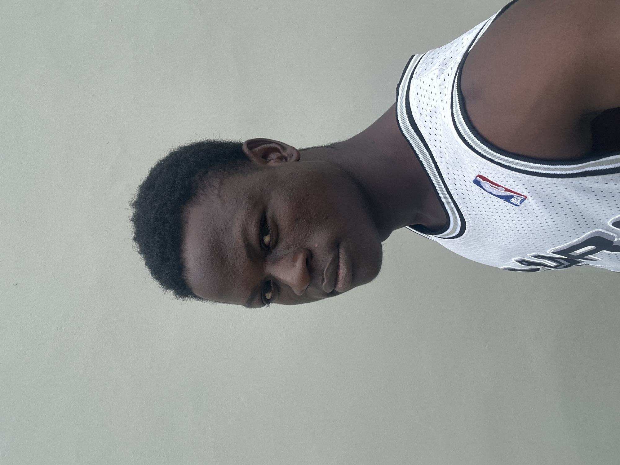 A close-up, waist-up, profile shot shows a young Black male with dark skin and short, dark, curly hair. He is wearing a white basketball jersey with black and silver trim, and the NBA logo is visible on the left side of the chest. He is looking directly at the camera with a neutral expression. The background is a plain, light-colored wall, suggesting an outdoor setting in Badagry, Nigeria. The lighting is even, with no harsh shadows. The overall impression is a casual, straightforward portrait.