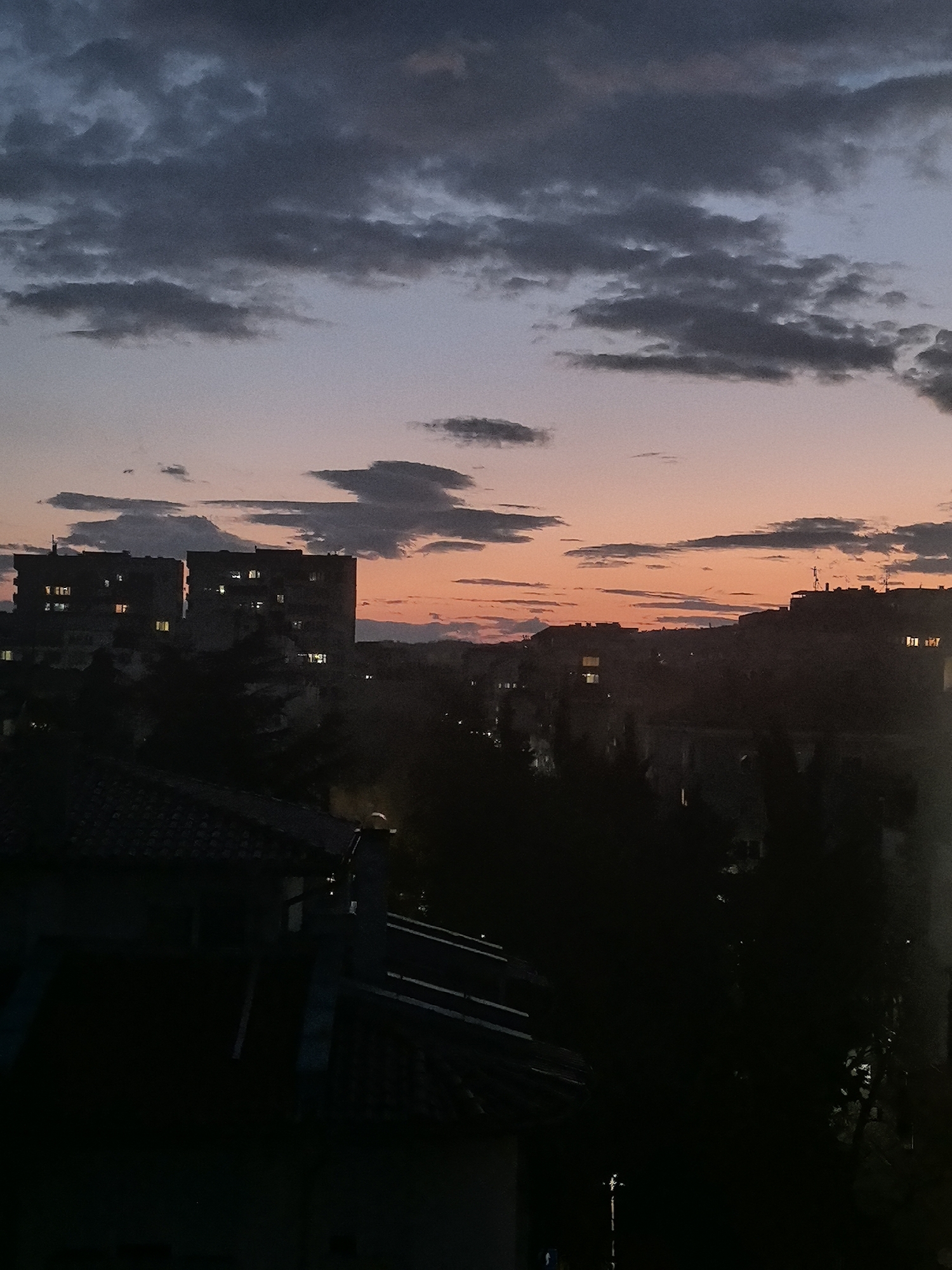 The image shows a cityscape at dusk or dawn, likely taken from an elevated position such as a balcony or high-rise building in Stara Zagora, Bulgaria. The sky is filled with dark grey and orange clouds, suggesting a sunset or sunrise. Several multistory apartment buildings are visible in the middle ground. Some windows are illuminated, indicating activity within. Dark outlines of rooftops and foliage occupy the foreground, adding depth to the image. The overall environment appears urban.