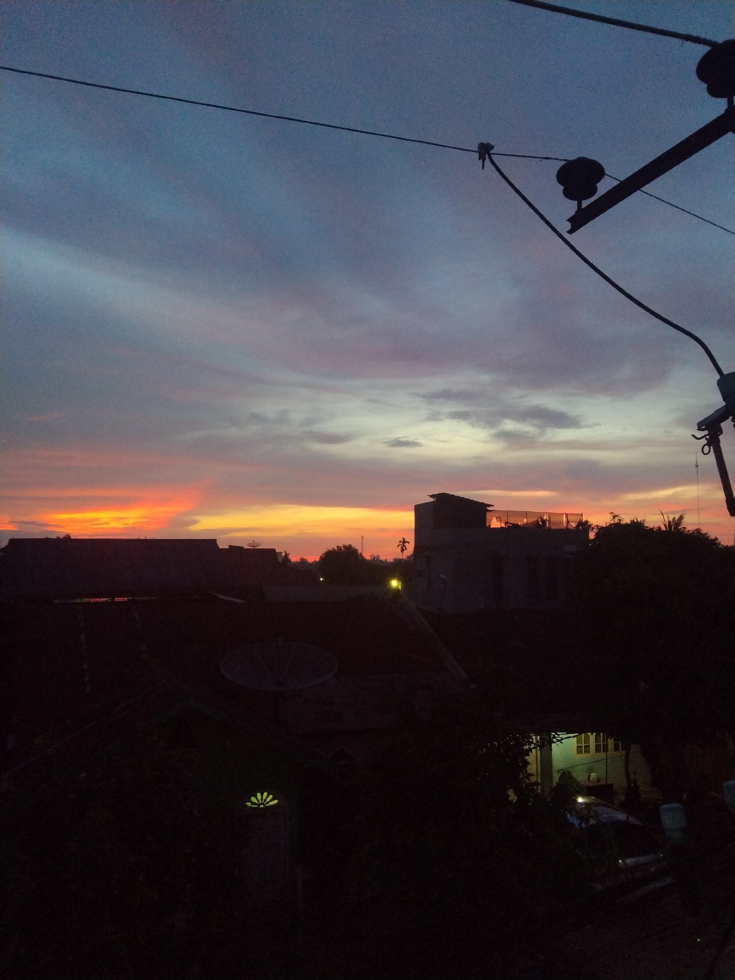 The image captures a twilight cityscape in Bireuenmeunasahdayah, Indonesia. The sky is a mix of blue, pink, and orange hues, suggesting either sunrise or sunset. Silhouetted rooftops dominate the foreground, one with a satellite dish. Electrical wires and a utility pole are visible in the upper right, adding to the urban feel. The buildings are dark and indistinct, with hints of foliage obscuring details. A faint glow emanates from a structure partially hidden by vegetation in the lower right corner. The overall impression is of a quiet, urban scene transitioning into night.