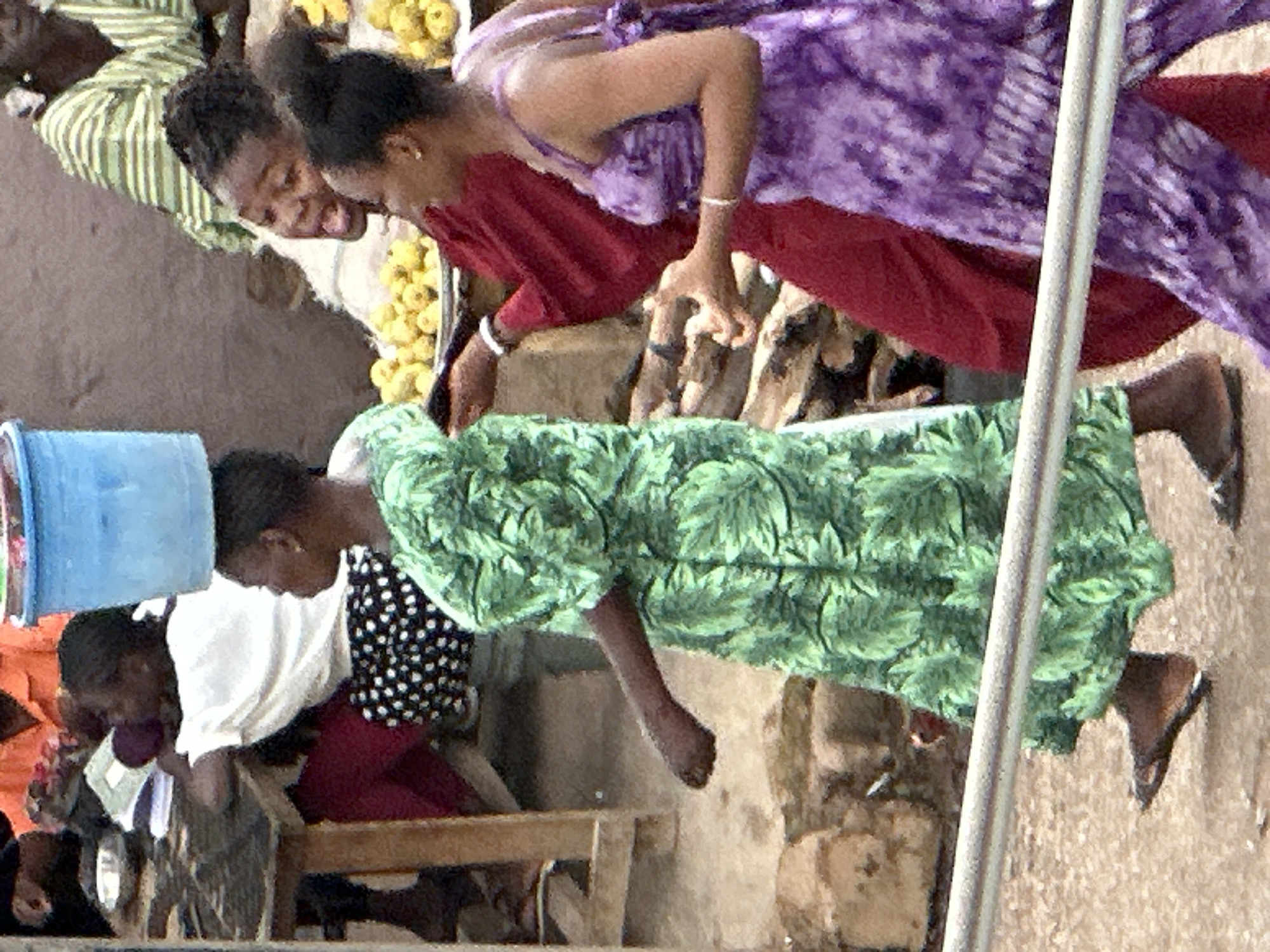 The image depicts an outdoor scene, likely a market, in Jos, Nigeria. In the foreground, a young woman wearing a vibrant green dress with a leaf pattern walks from left to right, carrying a large blue bucket on her head. Her left arm is extended outwards slightly as she walks. To her right, another woman is visible, wearing a red dress. Behind her, a woman in a purple tie-dye dress is partially visible. In the background, other individuals and market stalls can be seen. The ground appears to be dusty or dirt. The overall atmosphere suggests a busy commercial area.