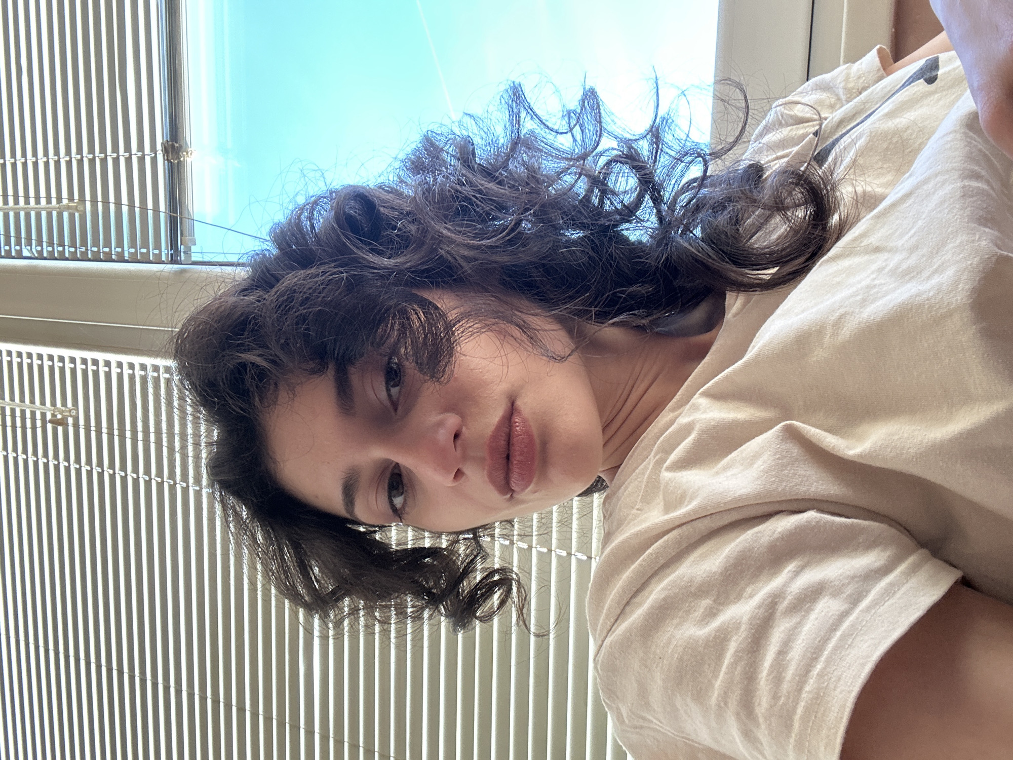 The image is a close-up selfie of a young woman with dark, curly hair and light skin. She is wearing a light-colored t-shirt. The background shows a window with horizontal blinds and a bright blue sky. The scene appears to be indoors, possibly in an apartment or office, with natural light coming through the window. The image does not contain any recognizable landmarks or specific architectural features that would allow for precise identification of a location within Sofia, Bulgaria.