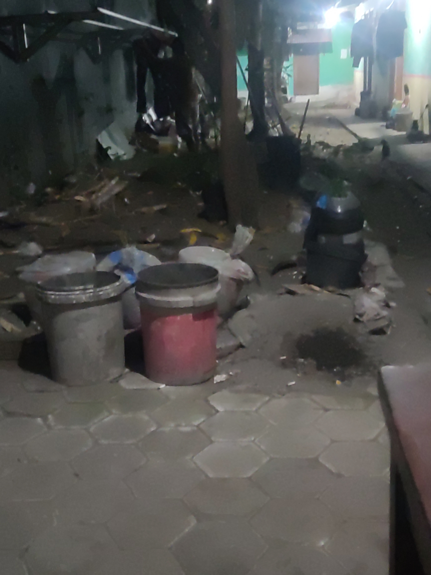 The image depicts a dimly lit outdoor scene in Trihanggo, Indonesia, likely in the evening or at night. In the foreground, a paved area with hexagonal stone tiles is visible. To the left, two large buckets or bins are situated on the tiles. One is a dark gray cylindrical bin, and to its right is a red cylindrical bin. Behind these bins, several large plastic bags, likely containing trash or refuse, are piled up.
The background reveals a cluttered and somewhat dilapidated environment. A low wall or structure runs along the left side, with some debris and wooden planks scattered near it. Further back, trees are present, casting shadows. Beyond the trees, buildings with simple facades are visible, one appearing to be painted green. Clothes are hanging from a line in front of one of the buildings. The overall impression is one of an unkempt urban or semi-urban area, possibly a residential or commercial back alley. No people are clearly discernible in the image, though shadows and indistinct figures may be present in the background. The lighting suggests artificial sources, likely from nearby homes or streetlights, creating a subdued and somewhat eerie atmosphere. The scene appears to be a collection point for trash or a general area of disarray.