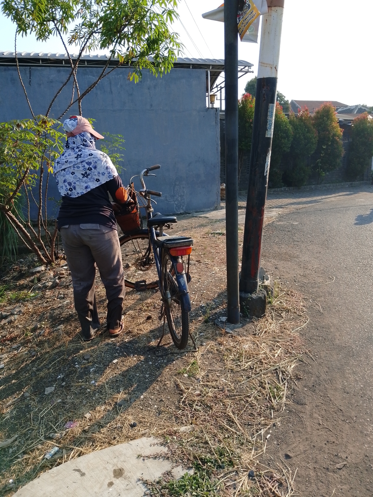 A person wearing a floral hijab, a dark long-sleeved shirt, and gray pants stands next to a bicycle on a street in Pasuruan, Indonesia. The person is facing away from the camera, holding the bicycle's handlebars. The bicycle has a basket on the front and a red taillight. The ground is covered in dry grass and dirt. A blue wall is visible behind the person and the bicycle. There is a utility pole with signs next to the bicycle. In the background, there are houses with red tile roofs and green trees. The sun is shining, casting shadows on the ground.