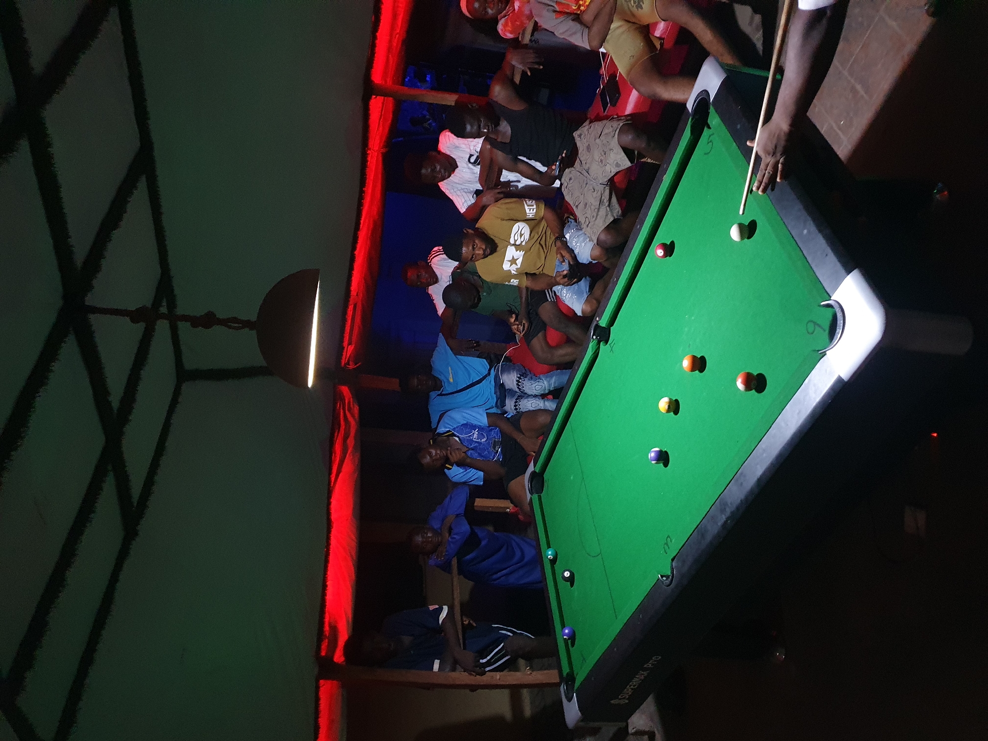 This photograph captures a lively evening scene of men gathered around a pool table, likely in a bar or social club in Lokoja, Nigeria. The setting is dimly lit, with prominent red lighting strips casting a vibrant glow along the walls and ceiling, creating a moody atmosphere.
The central subject is a green-felt pool table, on which several colored pool balls and a cue ball are scattered. One man is actively engaged in the game, leaning over the table with a pool cue in his hands, preparing to strike the cue ball. Other men are positioned around the table, some standing and watching the game intently, while others are seated on low benches or platforms along the perimeter of the room, socializing and observing. Their attire is casual, consisting of t-shirts, shorts, and jeans.
Notable details include a hanging light fixture visible in the upper left quadrant of the image, contributing to the overall illumination. The pool table itself is relatively well-lit, highlighting the game. The faces of many individuals are visible, suggesting a friendly and engaging social gathering. The image is taken from a slightly elevated perspective, looking down on the scene.
Visible text includes the brand name "SUPERMAX PRO" printed on the side of the pool table. The numbers "5" and "6" are faintly discernible on the white trim near the pockets of the table. The overall impression is one of casual leisure and camaraderie among friends or acquaintances.