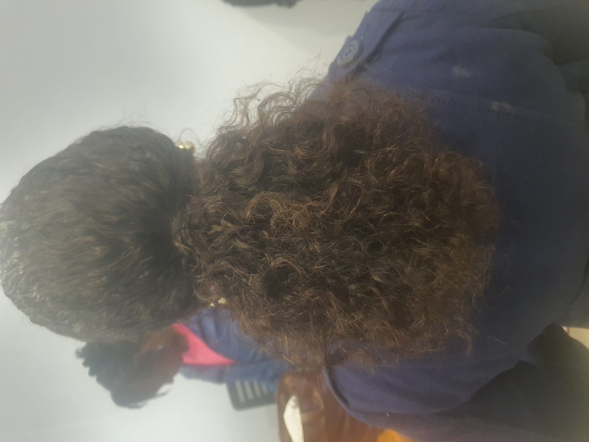 This media file is a close-up, overhead shot of the back of a person's head, showing their dark, curly hair. They are wearing a dark blue jacket with a button visible on the upper right portion of the image. A hint of a bright pink garment is visible underneath the jacket, near the person's shoulder. The lighting suggests an indoor setting, possibly with artificial light. The image appears to be taken in Jos, Nigeria, but no specific location cues are visible in the frame itself. The focus is entirely on the hair and the upper part of the jacket, giving no indication of the person's age, gender, or any particular activity or emotion. There is no visible text in the image.