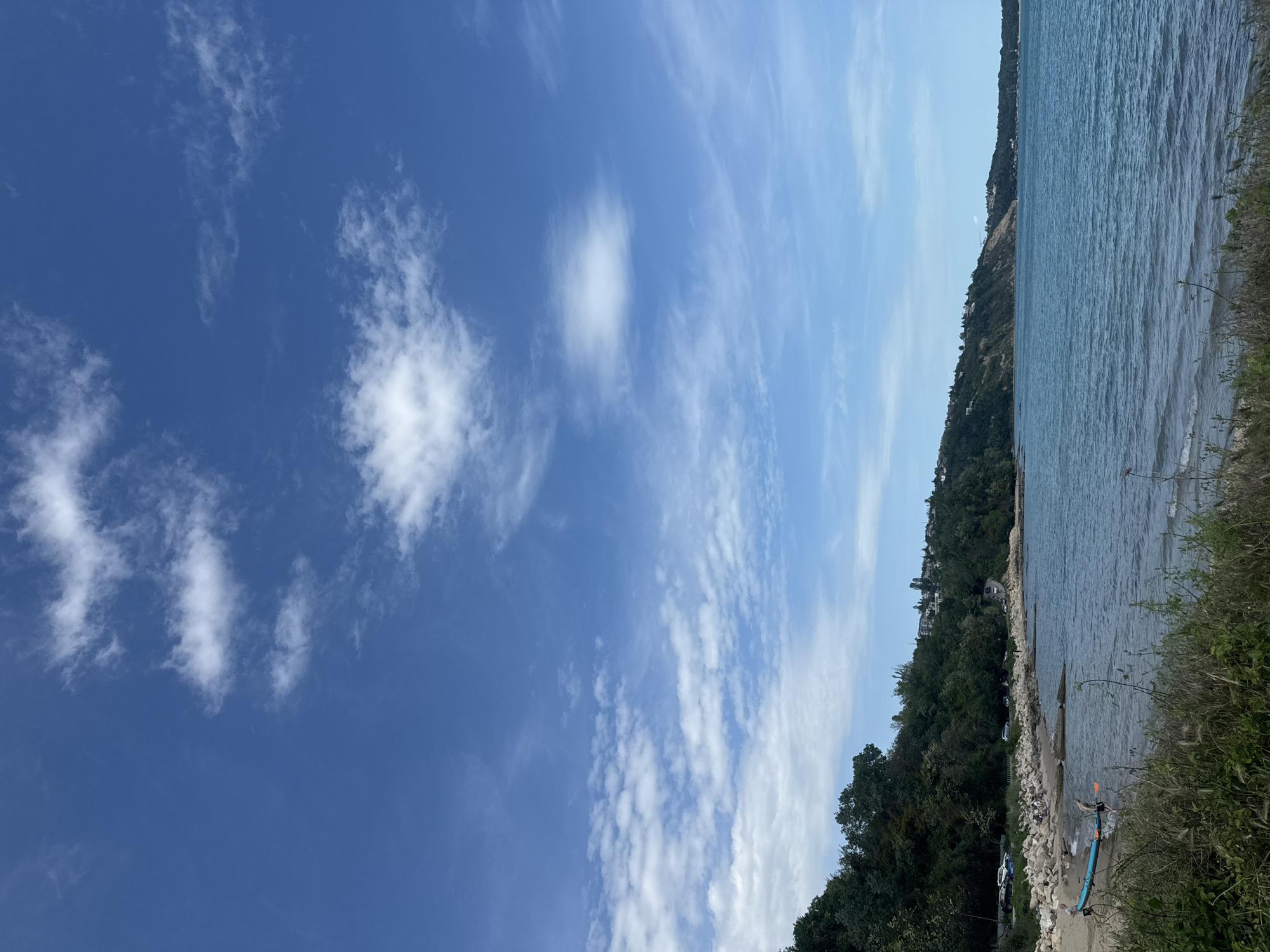 The image captures a scene in Varna, Bulgaria, featuring a view of the Black Sea. A rocky beach is visible, bordered by lush green vegetation and trees along a hillside. The sea is calm with gentle waves. In the lower right corner, there is a partially visible blue kayak. The sky above is mostly clear with scattered white clouds. The overall atmosphere suggests a sunny day near the coastline of Varna.