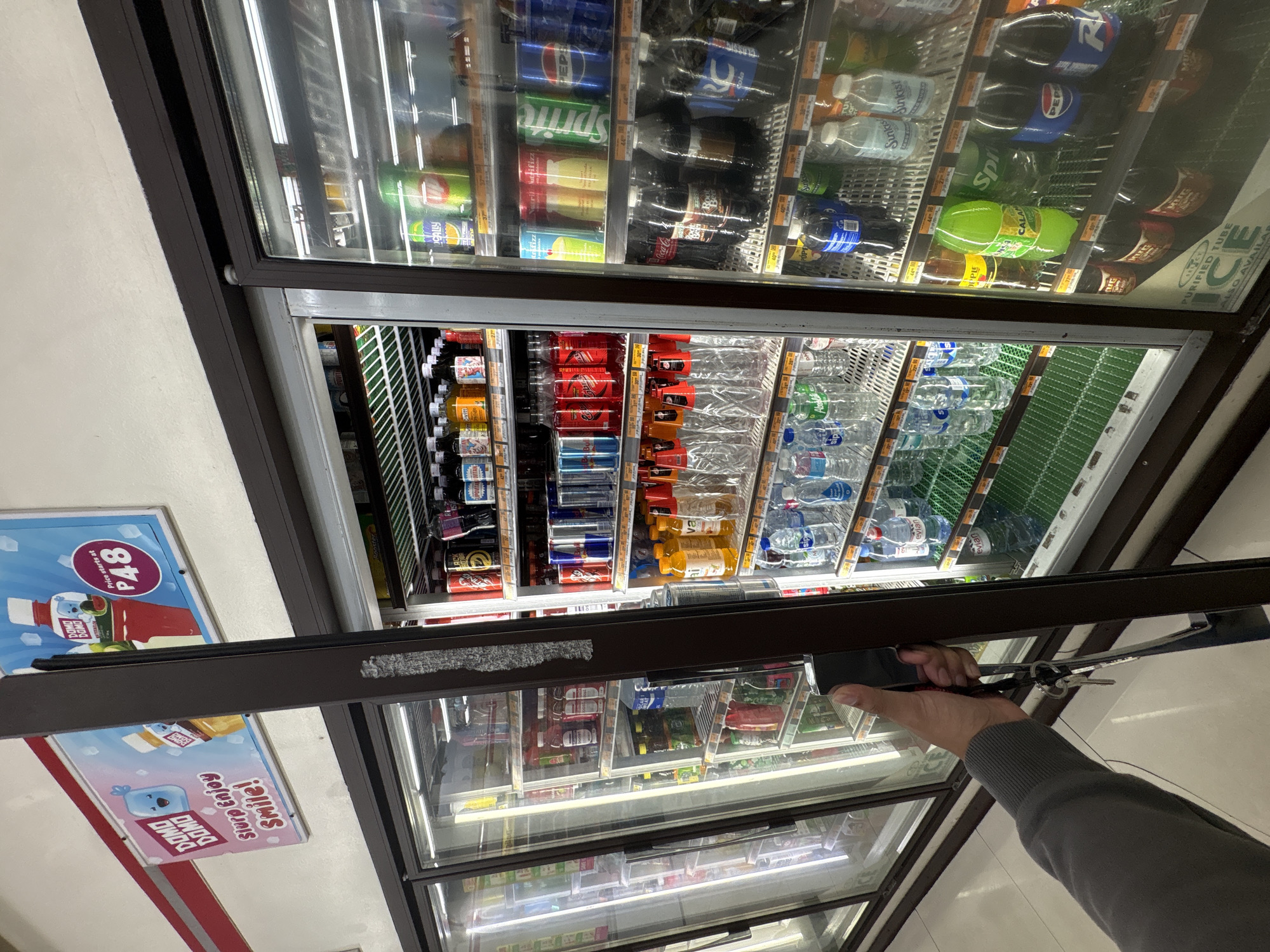 The scene depicts the interior of a convenience store, specifically focusing on a row of glass-front refrigeration units holding various beverages. In the foreground, a person's right hand, clad in a grey long-sleeved garment, is gripping the dark brown vertical handle of one of the refrigerators, which is slightly ajar. A set of keys attached to a black lanyard hangs from the handle area.
The refrigerators are brightly lit internally by fluorescent lights, illuminating shelves stocked with an assortment of canned and bottled drinks. The middle refrigerator, being opened, displays multiple tiers of products, including energy drinks (e.g., Red Bull, Black Mamba), various bottled juices, sodas (e.g., Pepsi, Sprite), and bottled water (e.g., Sip, Alps, Evian). Some empty plastic bottles are also visible on shelves. The refrigerator to the far right contains similar items, with recognizable brands such as Locally, Sunkist, Mug, Calamansi, Coca-Cola, Pepsi, and RC Cola. A sign advertising "Purified Tube Ice" is visible at the bottom of this unit.
Above the refrigerators, on a white wall, two rectangular signs are mounted. The left sign features a blue, smiling, blob-like cartoon character with the text "Domo Domo Slurp Enjoy Smile!". The adjacent sign displays a bottle labeled "Domo Domo Watermelon" with "Price starts at P48" indicated, suggesting pricing in Philippine Pesos. The store's ceiling is white, and the floor is composed of light-colored tiles with darker grout lines, visible in the lower portion of the image. The entire scene is consistent with a commercial establishment, likely a 7-Eleven or similar convenience store, located in Davao City, Philippines.