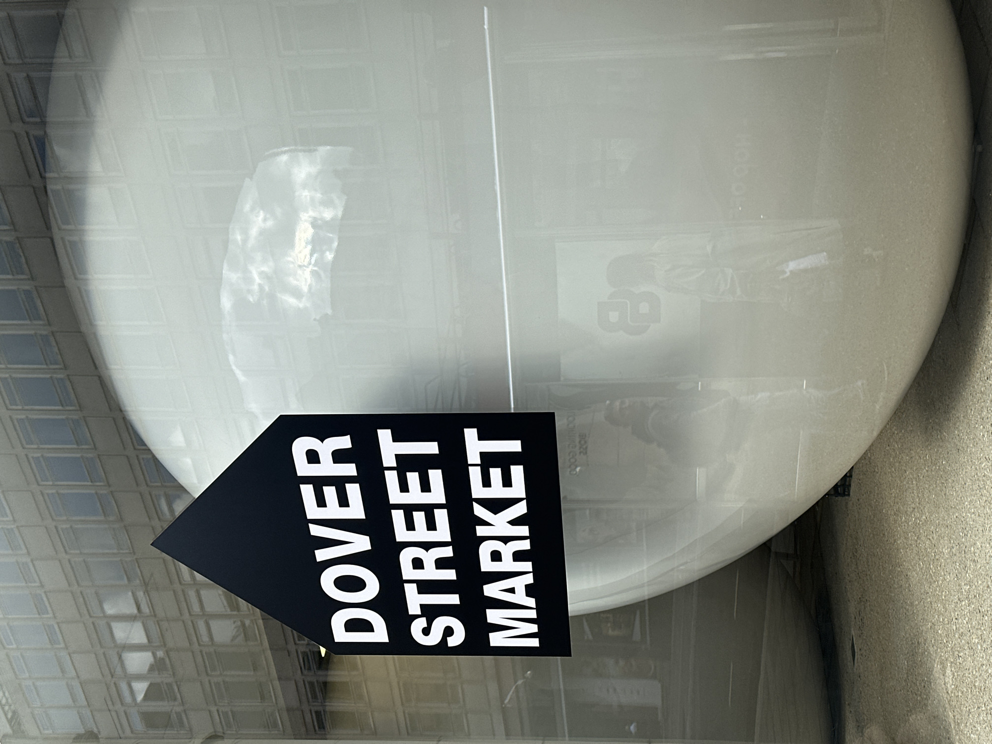 The image shows a black arrow-shaped sign with "DOVER STREET MARKET" printed in white, positioned in front of a window display. The display features a large, rounded, light-colored object, possibly an oversized sculpture or decoration. Reflections in the window reveal an exterior view of a building with a grid-like pattern of windows, suggesting an urban setting. The presence of "Dover Street Market" on the sign indicates that the location is likely in London, United Kingdom, specifically in or near Dover Street, known for its high-end fashion and retail establishments. The scene appears to be an exterior shot taken during the day, with natural light illuminating the display.