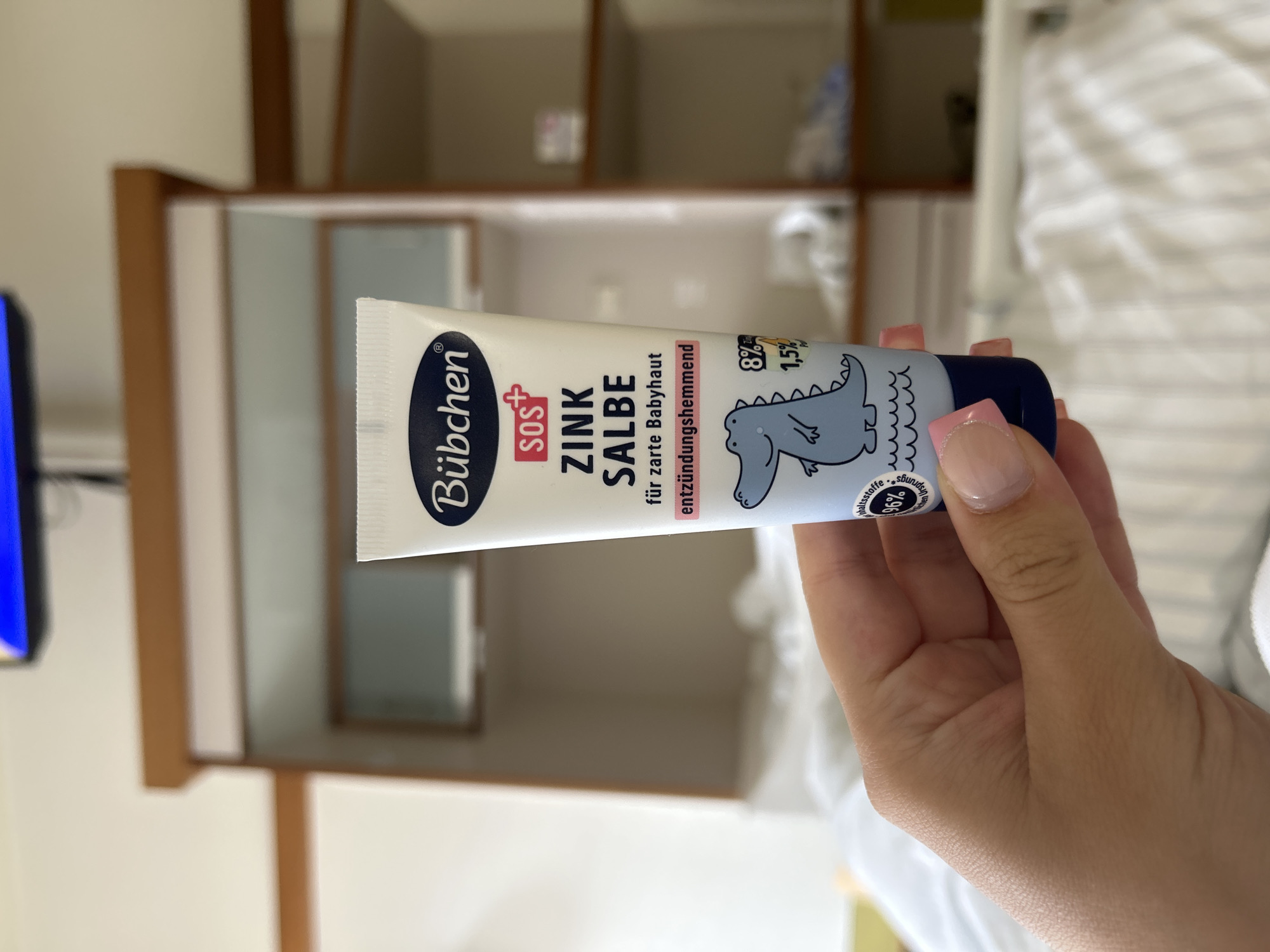 A person's hand, with a light pink manicure, holds a tube of "Bübchen SOS Zink Salbe" cream in front of a blurred background. The background appears to be an indoor setting, possibly a hotel room or hospital room, with a bed and shelves visible. The tube of cream features a blue and white design with an illustration of a dinosaur. The text on the tube indicates it is a zinc ointment for sensitive baby skin and has anti-inflammatory properties. The lighting is natural, suggesting daylight. The scene is photographed from a slightly elevated angle, focusing on the hand and the cream.