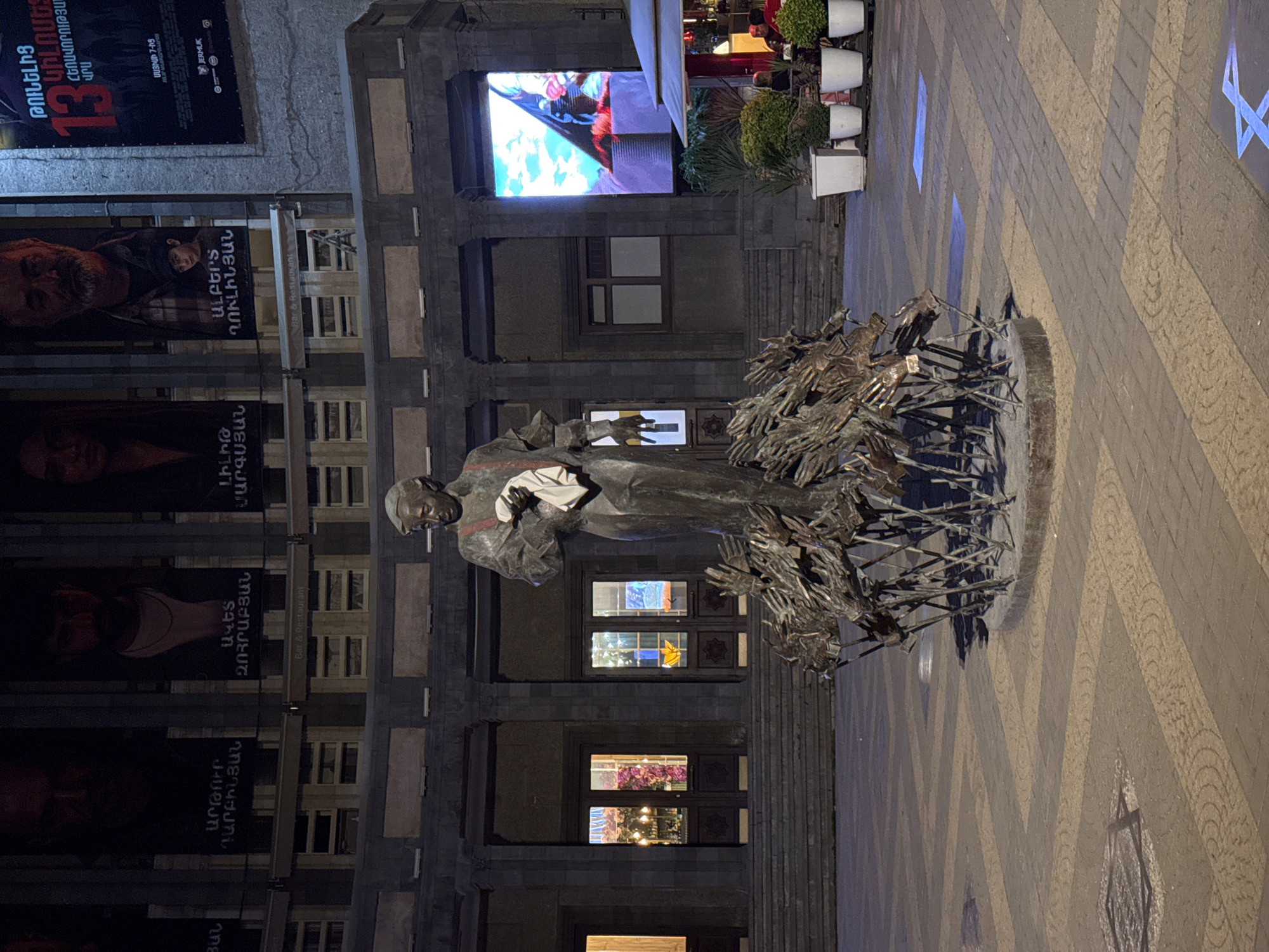The image shows a statue in Yerevan, Armenia, at night. The statue is of a figure holding a book, surrounded by a structure of metal arms. Behind the statue is a building with windows and posters of a film titled "ԹՈՒՆԵԼԻՑ 13," featuring the names of Armenian actors. A large screen displays colors on the building's upper level. The ground is paved with a geometric pattern. Plant pots line the perimeter of the upper building level.