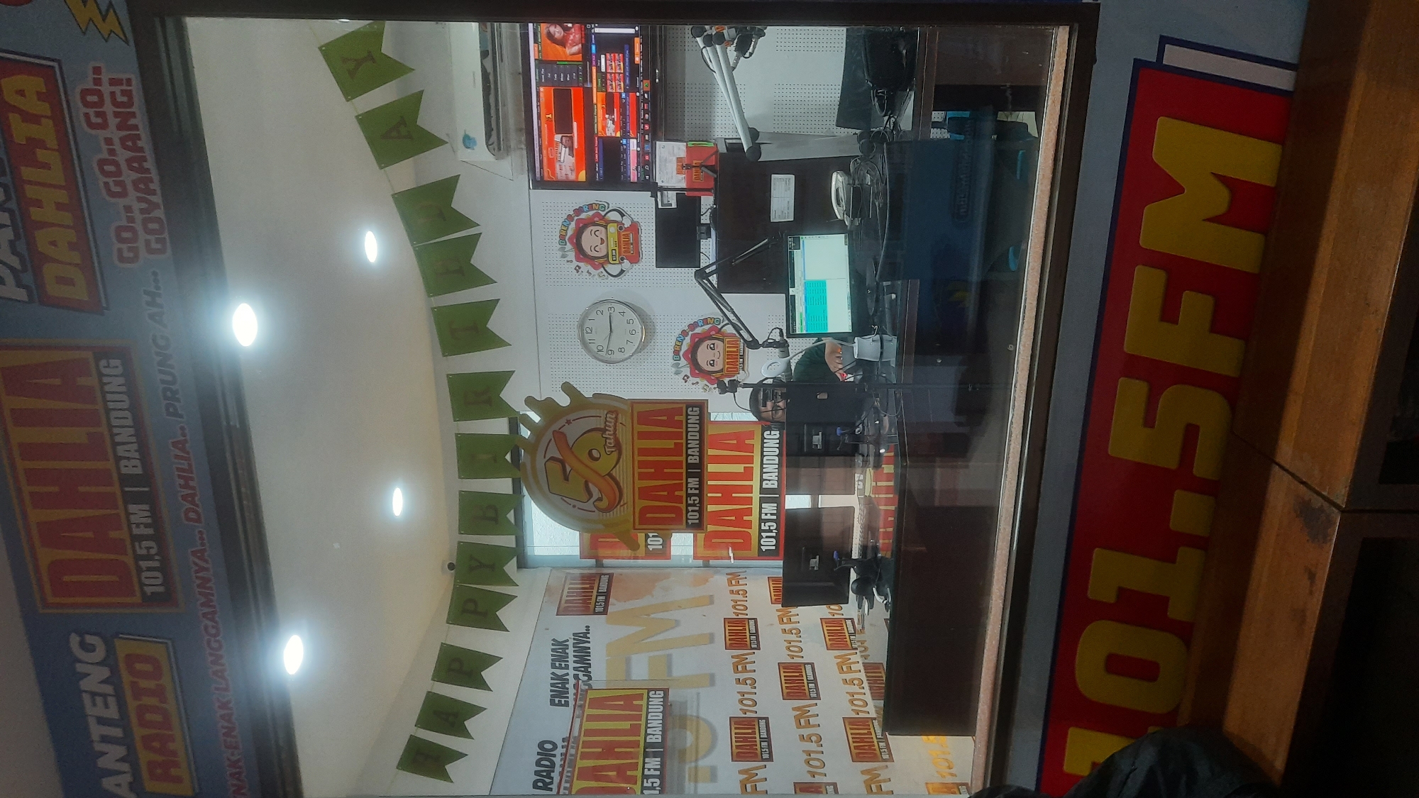 The image shows the interior of a radio station, "DAHLIA 101.5 FM BANDUNG," decorated for a celebration. A banner reading "HAPPY BIRTHDAY" hangs across the ceiling. There are several prominent signs displaying the radio station's logo and frequency. Inside the studio, a person wearing headphones is seated at a desk with multiple computer monitors and audio equipment. A large clock on the wall indicates the time is approximately 12:40. The overall environment suggests a lively atmosphere within the radio station. The specific location within Bandung City is not discernible from the image itself.