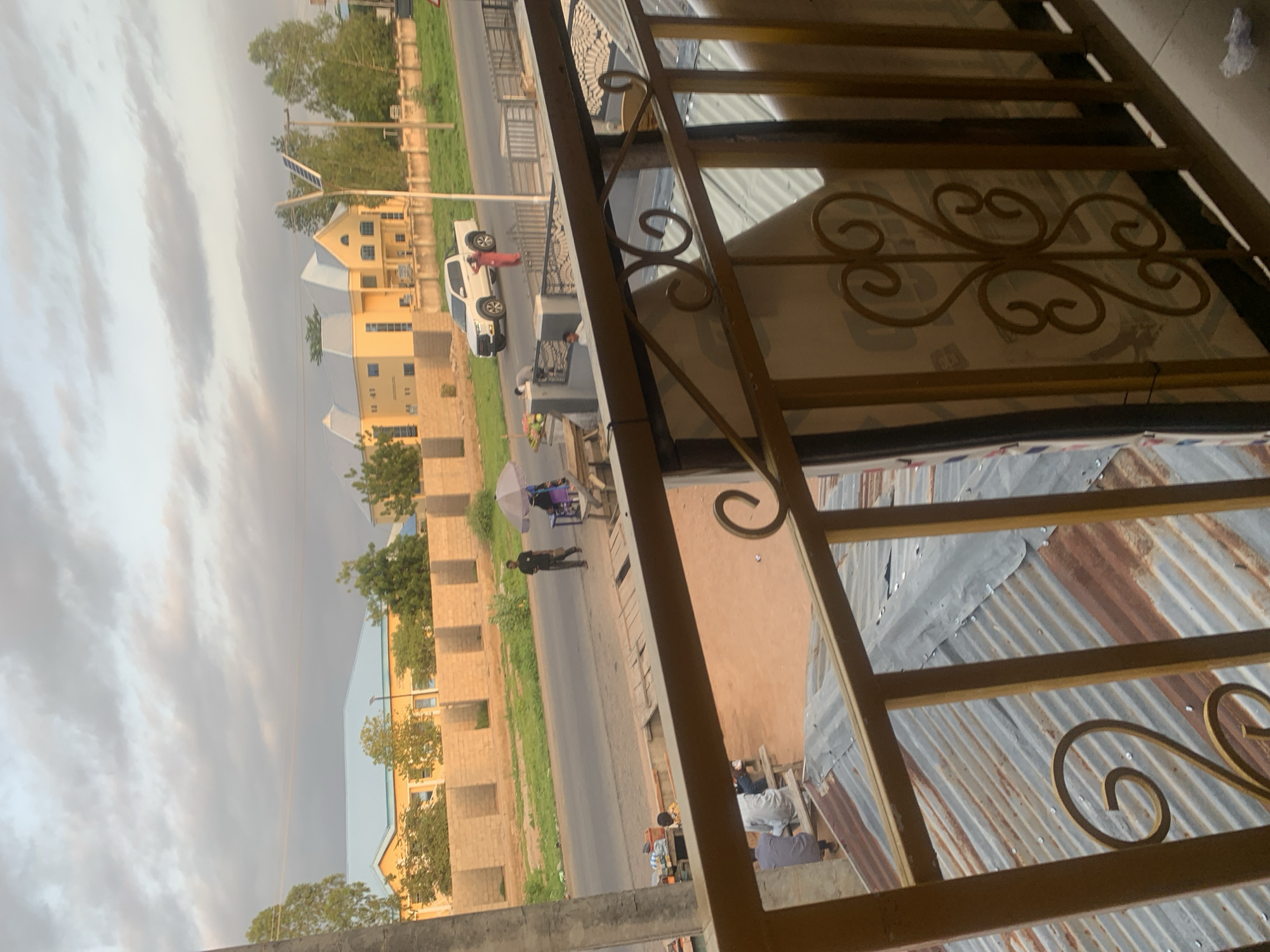The image depicts a street scene in Jalingo, Nigeria, under a cloudy sky. In the foreground, a rusted corrugated metal roof is visible, partially obscured by a golden metal railing with decorative scrollwork. Beyond the railing, a paved road runs horizontally across the frame. On the far side of the road, a low, rough-hewn brick wall separates the road from a grassy area and buildings.
Several people are present on or near the road. To the left, a person in a dark shirt stands near a table with what appears to be goods for sale, including some fruit like watermelons. Another person is seated at a table further left. In the middle ground, two figures in dark clothing walk along the road. To the right of the road, a white pickup truck is parked, and a person in a red garment stands nearby, facing away from the camera. Further down the road, there are more buildings. One prominent building is light yellow with a light blue roof and geometric architectural features. Another building behind it is also yellow and has a distinctive roofline.
The environment appears to be a somewhat developed urban area, with paved roads, buildings, and a mix of people engaged in various activities, including what seems to be street vending and transportation. The overall lighting suggests it might be late afternoon or early evening.
