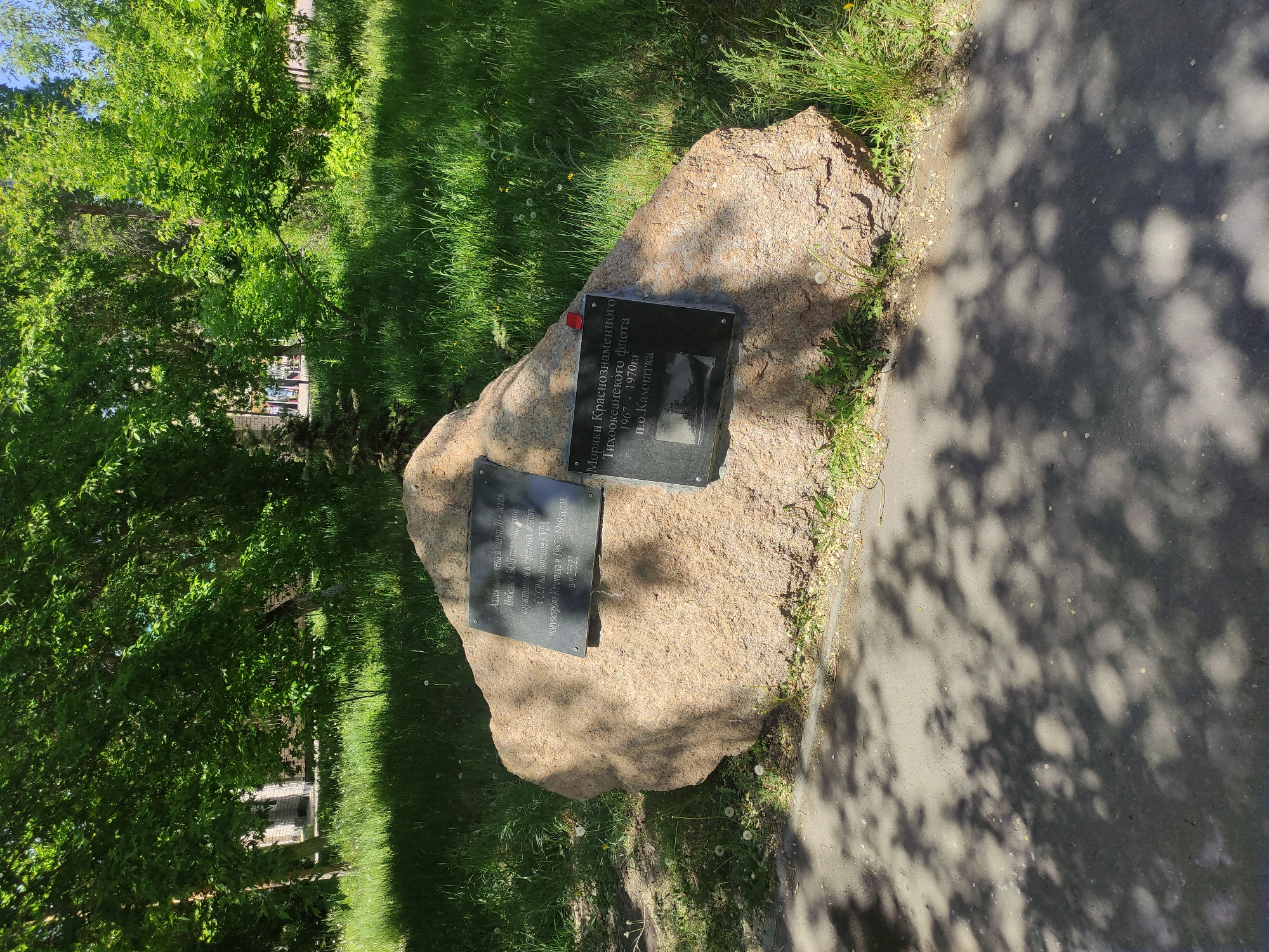 The image shows a memorial stone in Pavlodar, Kazakhstan, dedicated to veterans. Two plaques are affixed to the stone. The top plaque commemorates sailors of the Red Banner Pacific Fleet stationed on the Kamchatka Peninsula from 1967 to 1970. The bottom plaque commemorates Pavlodar residents who served in the USSR missile forces at the KURA test range on the Kamchatka peninsula from 1967-1969. The memorial is located in a green space, adjacent to a sidewalk. Trees and grass surround the stone.