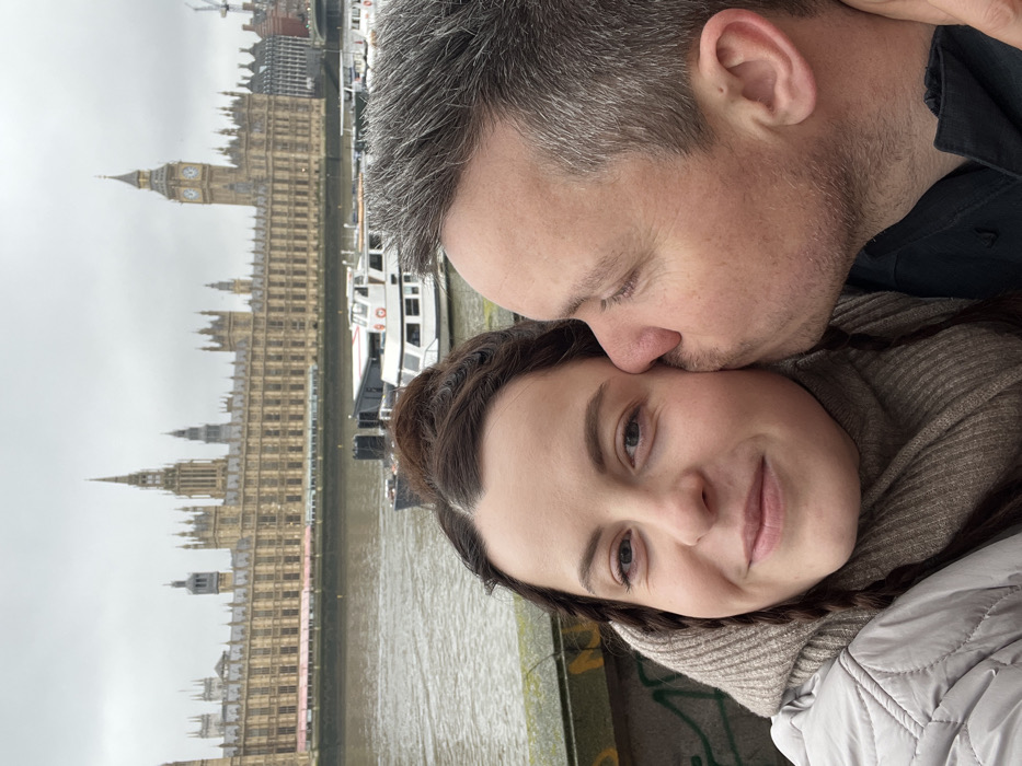 The image depicts two individuals in a close-up, outdoor shot against an overcast London backdrop. A woman with dark hair, styled with braids and a top bun, is on the left, looking directly at the camera with a subtle smile. She wears a light-colored puffer jacket and a ribbed beige scarf or hood. On the right, a man with short, grey-speckled hair is partially visible, kissing the woman's right cheek.
Behind the individuals, a moss-covered concrete or stone barrier suggests their position on a river embankment or bridge. In the mid-ground, across the River Thames, the Palace of Westminster (Houses of Parliament), including the Elizabeth Tower (Big Ben), is clearly visible under a grey sky. Several small to medium-sized vessels are docked or moving on the river between the foreground subjects and the iconic landmark.
