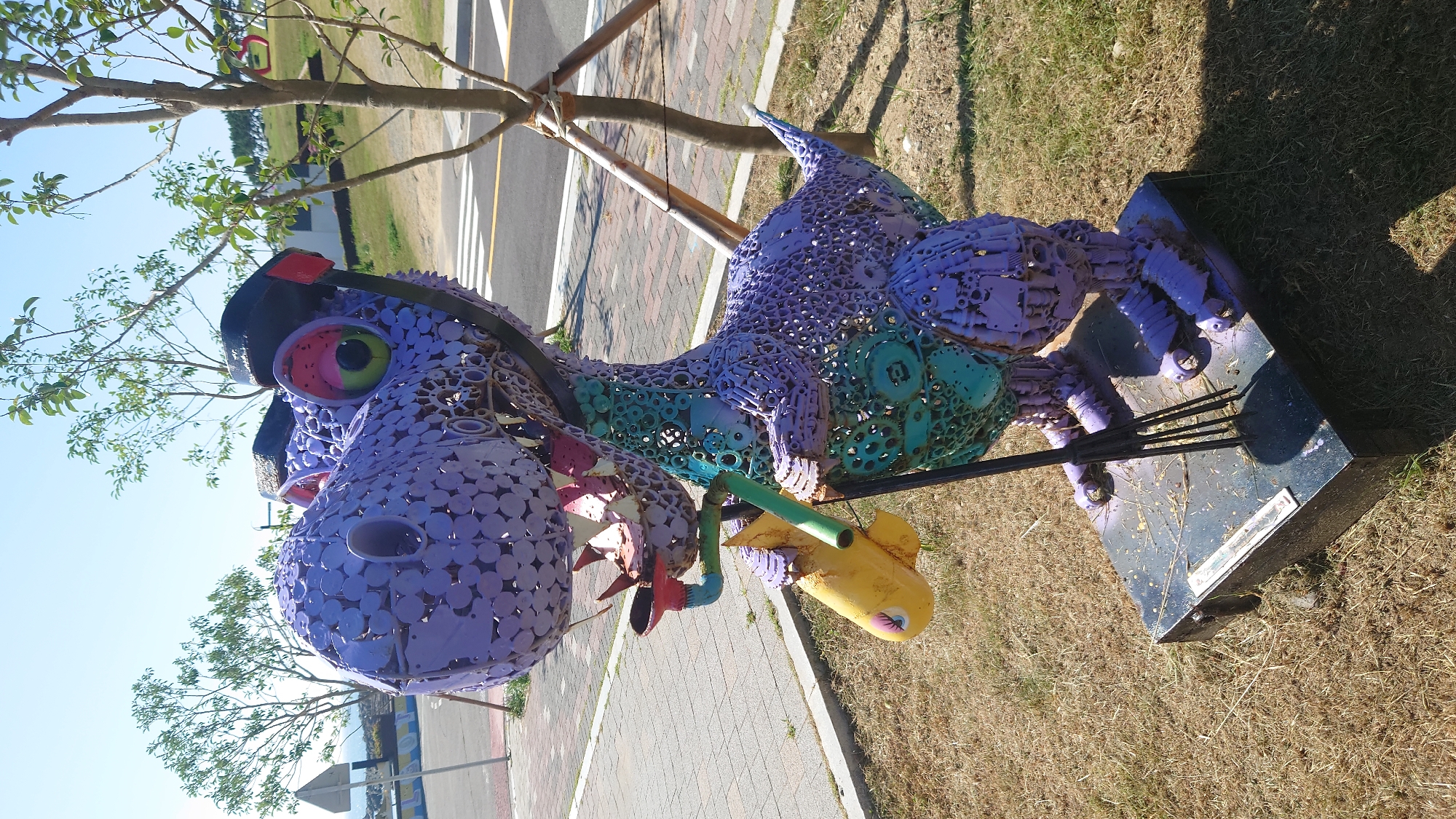 An outdoor sculpture of a purple, metallic dinosaur is displayed on a paved area next to a grassy field. The dinosaur is constructed from various metal pieces, including what appear to be gears and circular components, and is painted predominantly purple, with accents of teal on its chest and pink and green in its eyes. It wears a black headband and goggles. In its right hand, it holds a yellow object resembling a stylized banana, and its left hand clutches a black rake. The dinosaur stands on a dark-colored, rectangular base. Behind the sculpture, a paved pathway runs alongside a road with crosswalk markings. Trees are visible in the background, with a clear blue sky overhead. The location is the Bangudae Petroglyphs Museum in Ulsan, South Korea.