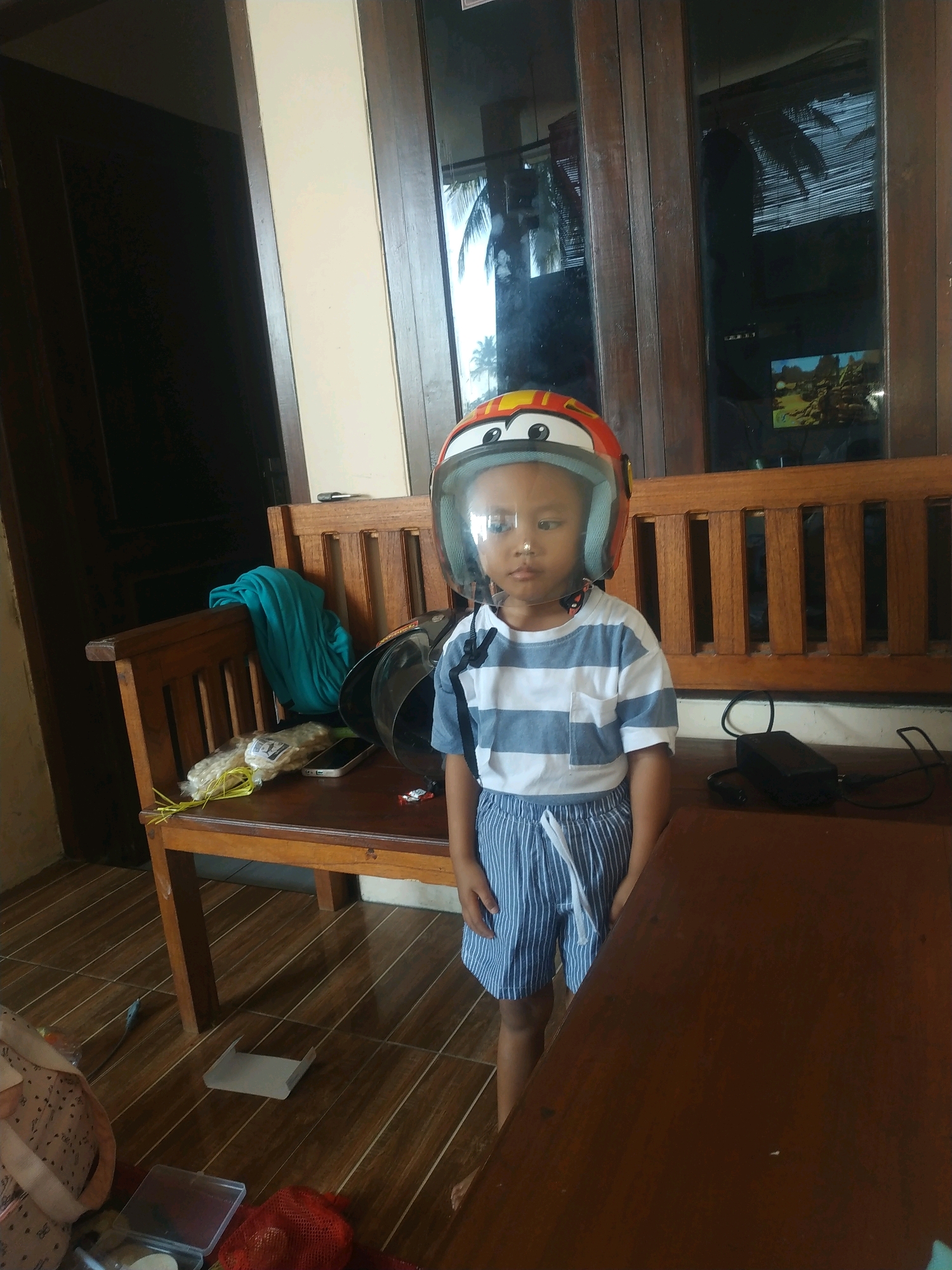 A young child wearing a cartoon-themed helmet stands on a tiled floor next to a wooden bench and table. The child is wearing a blue and white striped t-shirt and blue striped shorts. A teal fabric is draped over the back of the bench, and several other items, including a phone and a bag of yellow objects, are on the bench. A black electronic device with cords is on the table. Behind the child is a wooden structure with glass panes reflecting palm trees and a distant landscape. The scene appears to be indoors, possibly in a home or guesthouse, in a tropical environment, given the reflection in the glass. The overall impression is of a casual, everyday moment.