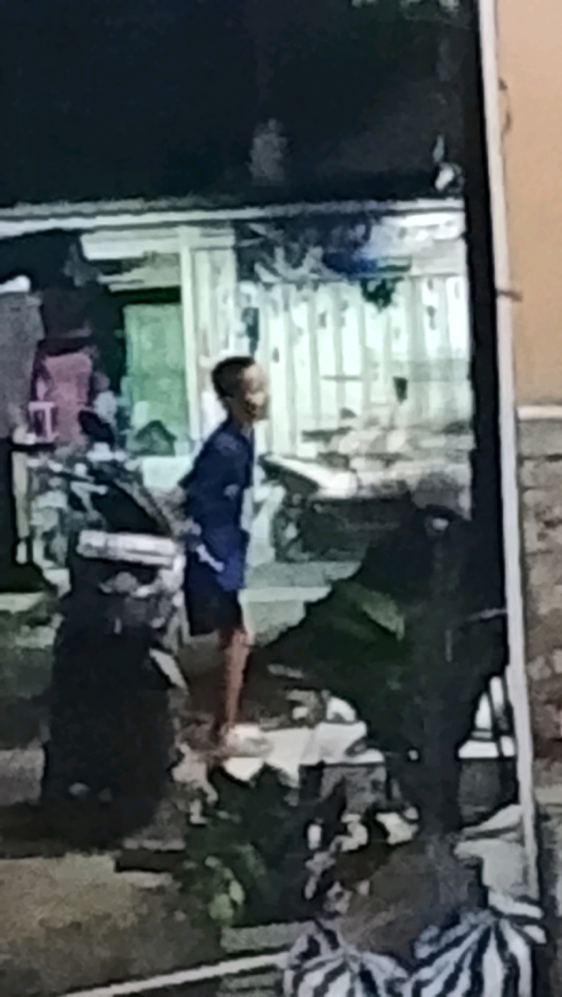 The image depicts a young male, appearing to be of Indonesian ethnicity, standing in what appears to be a back alley or an outdoor storage area. He is wearing a blue long-sleeved shirt and dark shorts. The background shows a cluttered environment with various objects, possibly including storage containers, debris, and some sort of shelter or structure with vertical elements. The overall lighting suggests it might be evening or a dimly lit area. There are no immediate interactions visible between the subject and other individuals. The location is Ujungrusi, Indonesia.