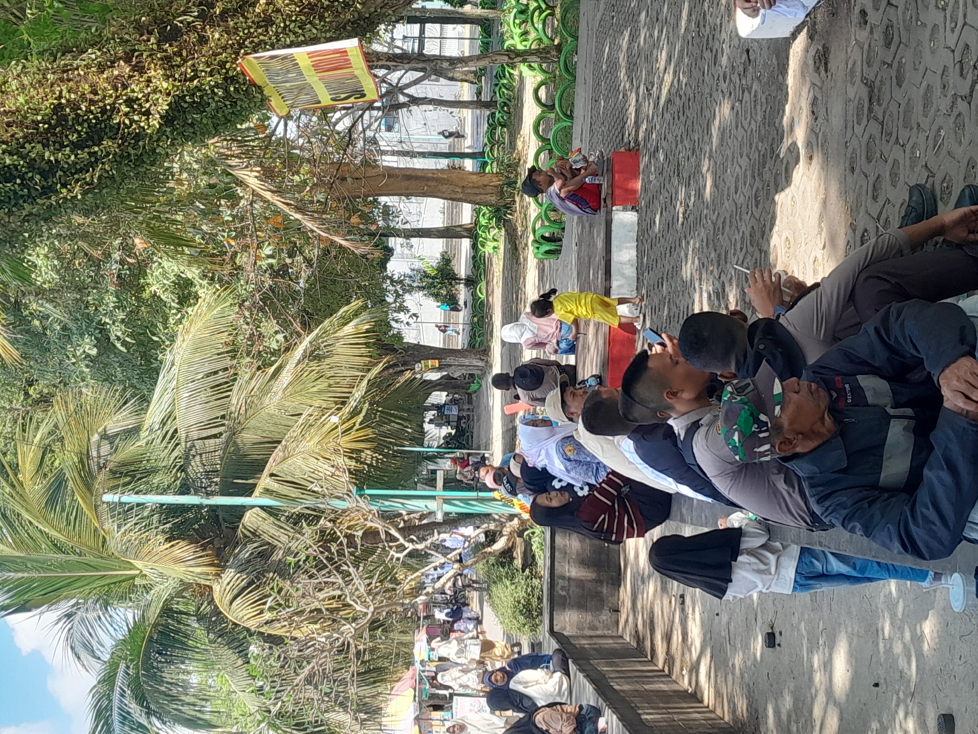 The image depicts a public area, likely a park or recreational space, in Genteng Kulon, Indonesia. Numerous individuals, predominantly seated on a concrete ledge or on the ground, are visible. The atmosphere appears relaxed, with some people conversing or observing their surroundings. A young child in a yellow outfit stands out, seemingly interacting with an adult. Palm trees and other vegetation are prominent, indicating a tropical or subtropical environment. In the background, a yellow banner with text is partially visible, and various structures suggest a developed public space. The ground is paved with interlocking stones.